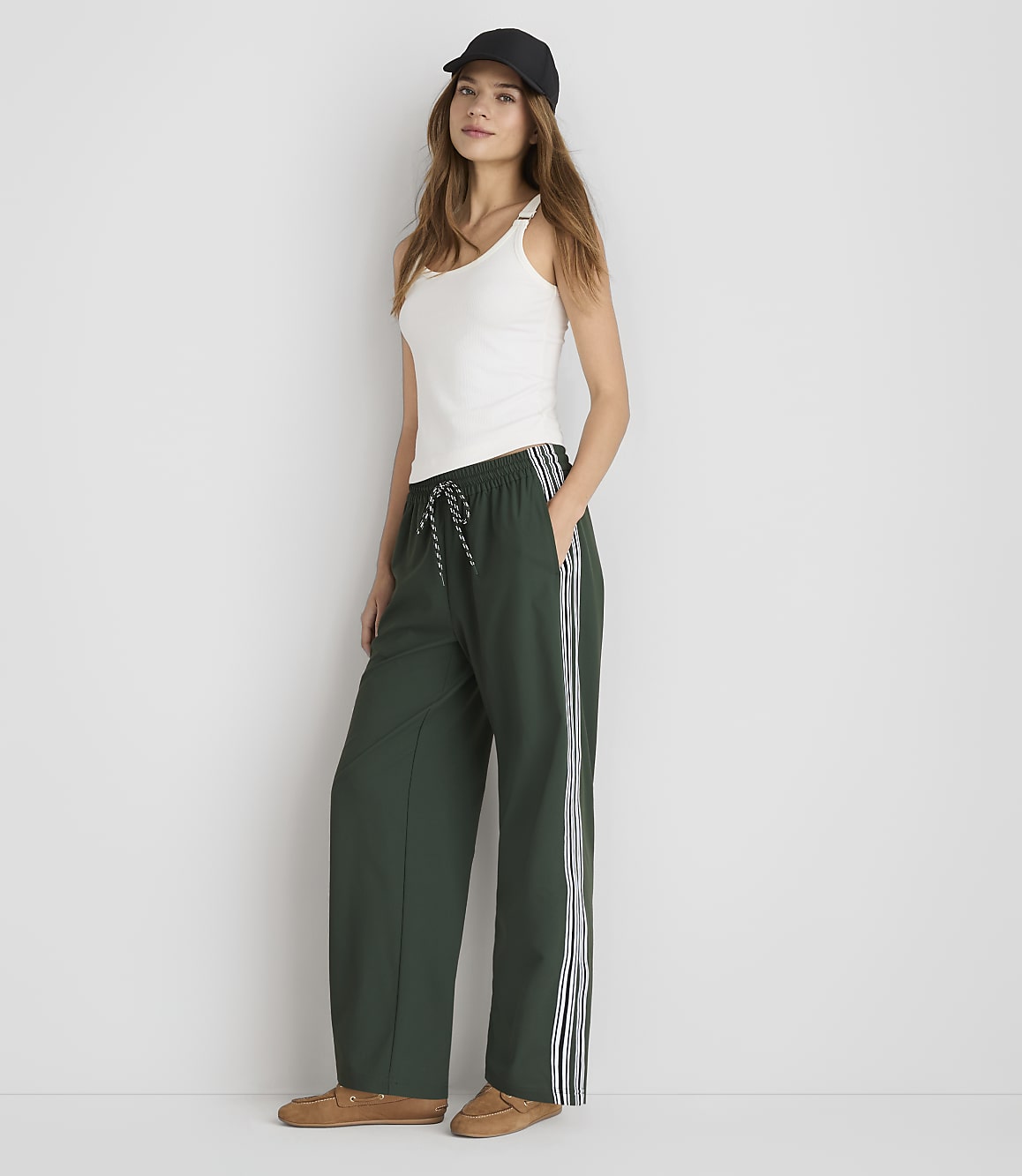 A woman in a white tank top and green track pants stands against a plain white background.
