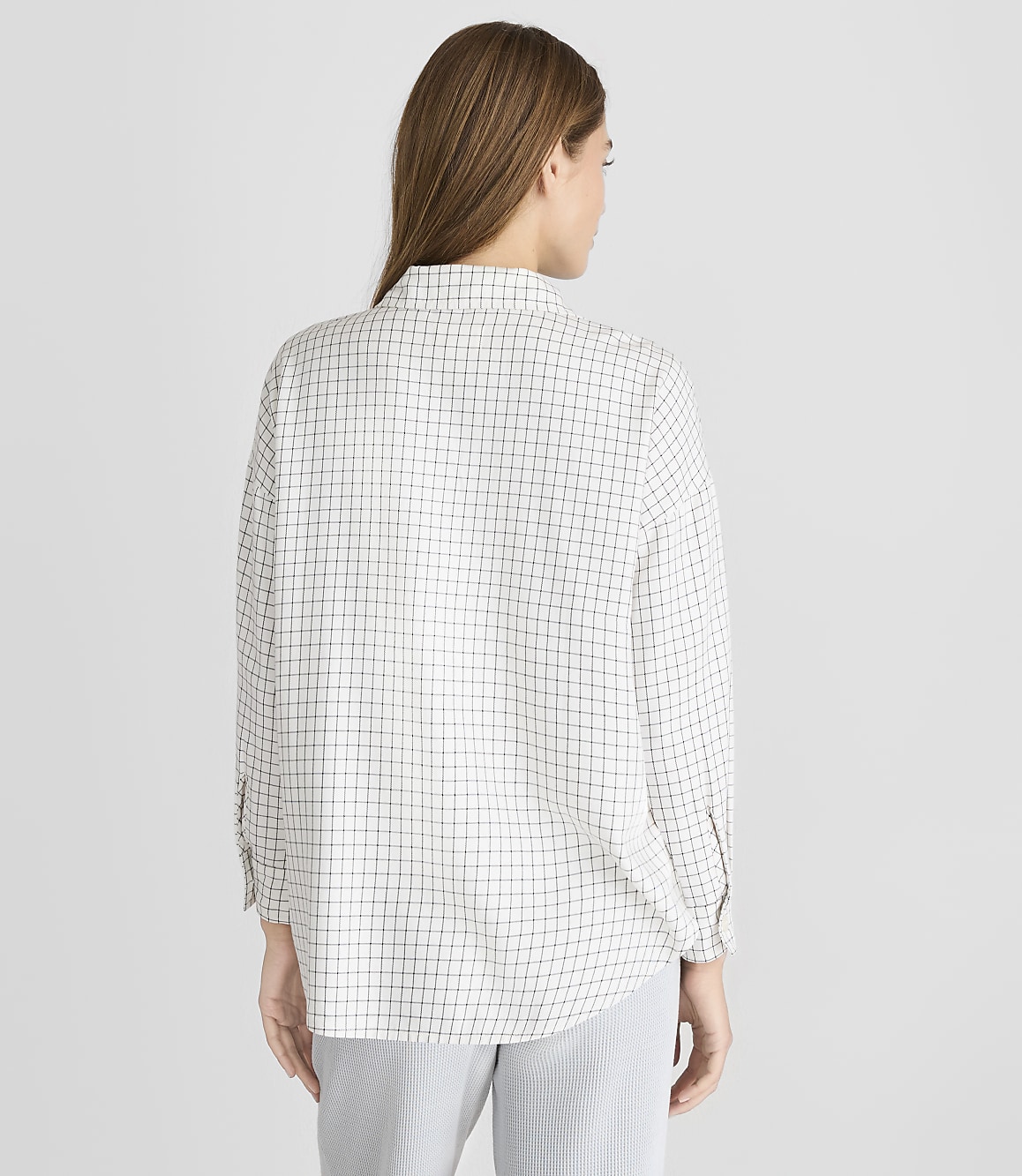 A woman wearing a checkered shirt and striped pants stands against a plain gray background.