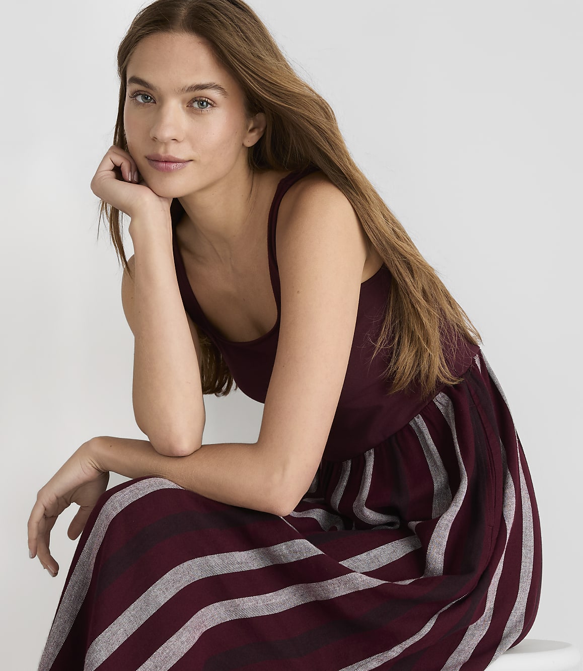 A young woman with long brown hair rests her chin on her hand, wearing a maroon and gray striped dress against a plain white background.
