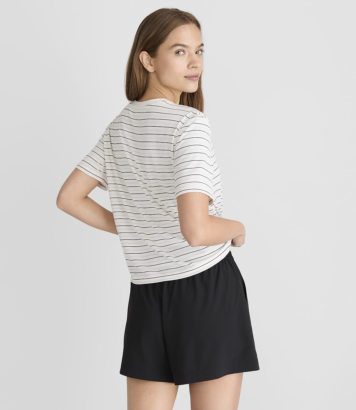 A young woman in a striped t-shirt and black shorts stands against a plain gray background.