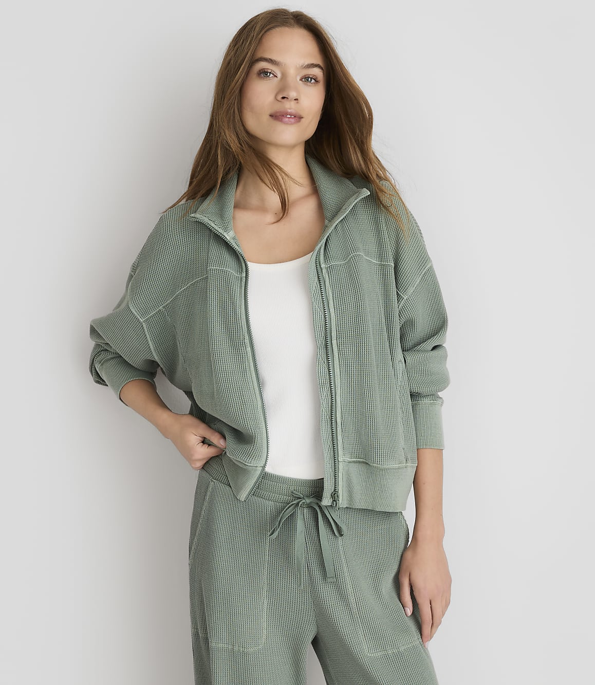 A woman in a green zip-up jacket and matching pants stands against a plain white background.