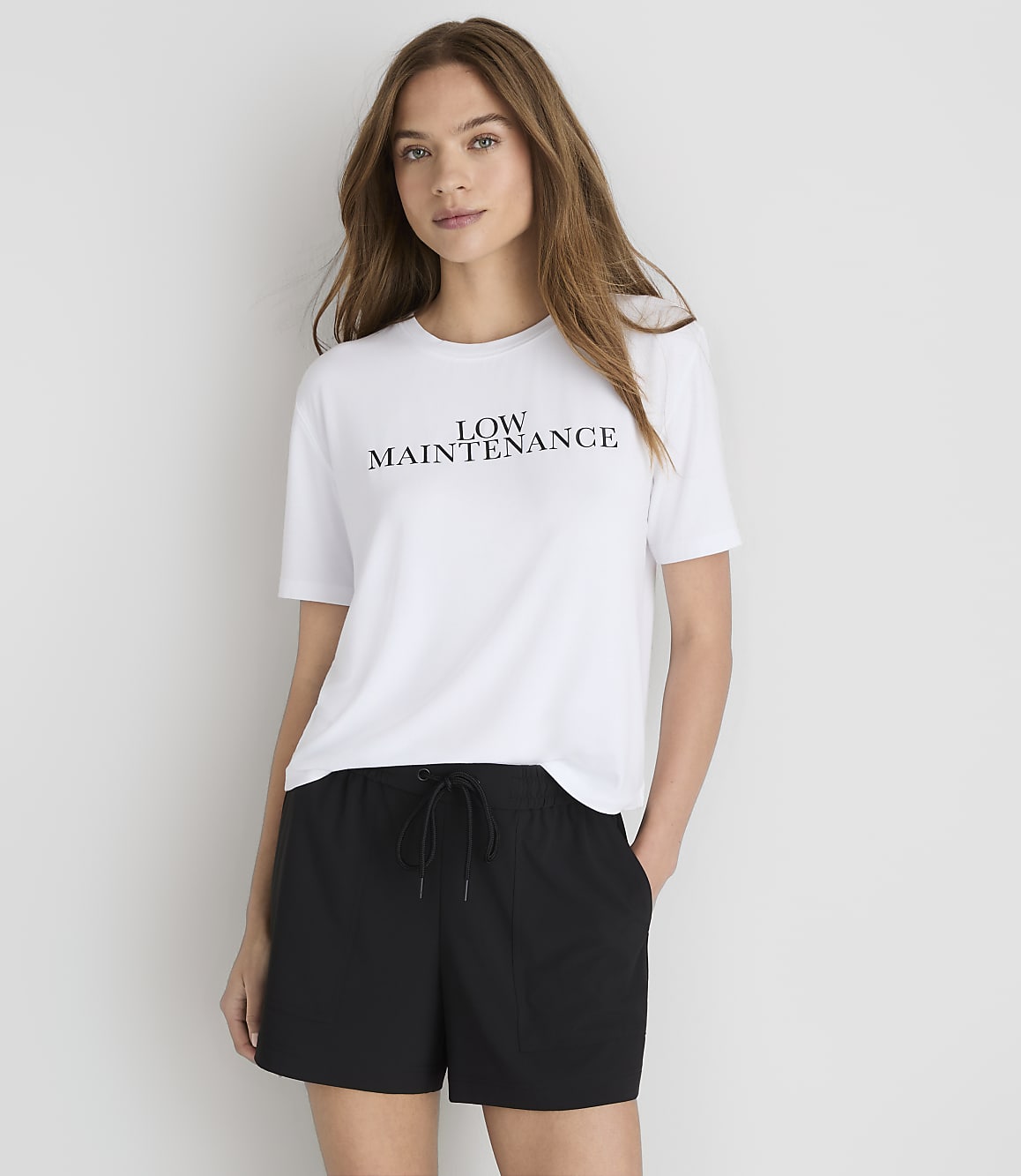 A woman in a white t-shirt with "LOW MAINTENANCE" printed on it and black shorts stands against a plain white background.