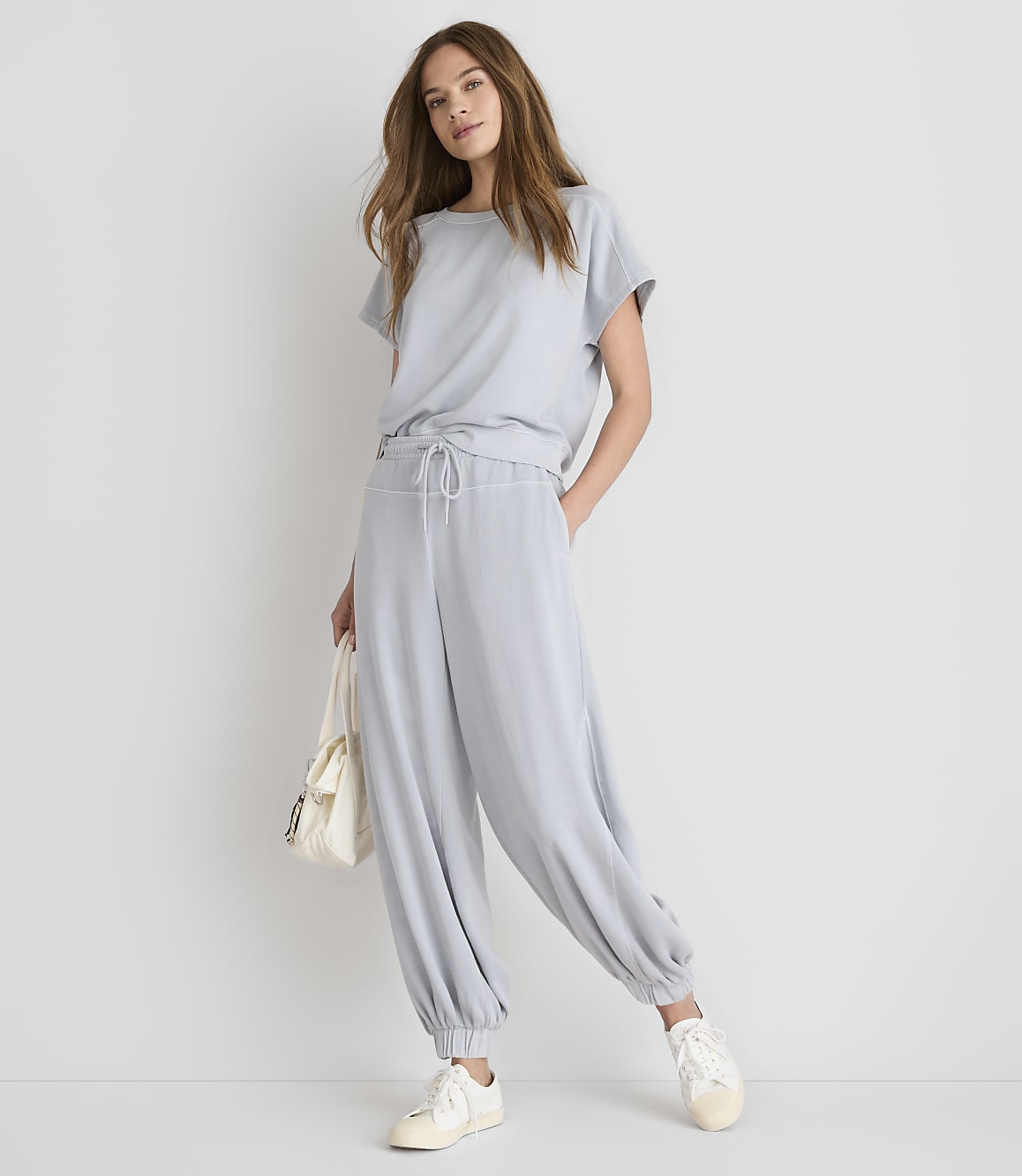 A woman in a light blue loungewear set and white sneakers stands against a plain white background, holding a cream-colored tote bag.