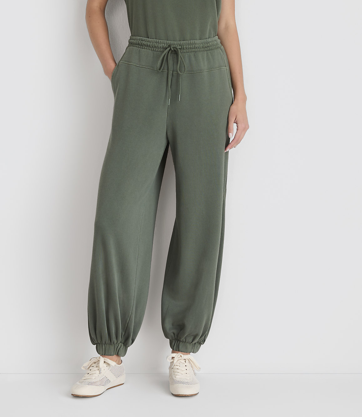 A person wearing olive green sweatpants and sneakers stands against a plain white background.