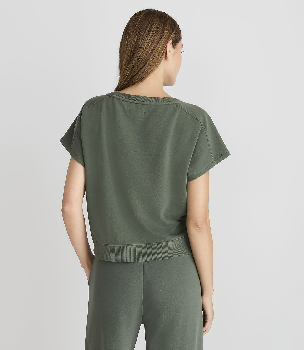 A woman in a green t-shirt and pants stands against a plain gray background.