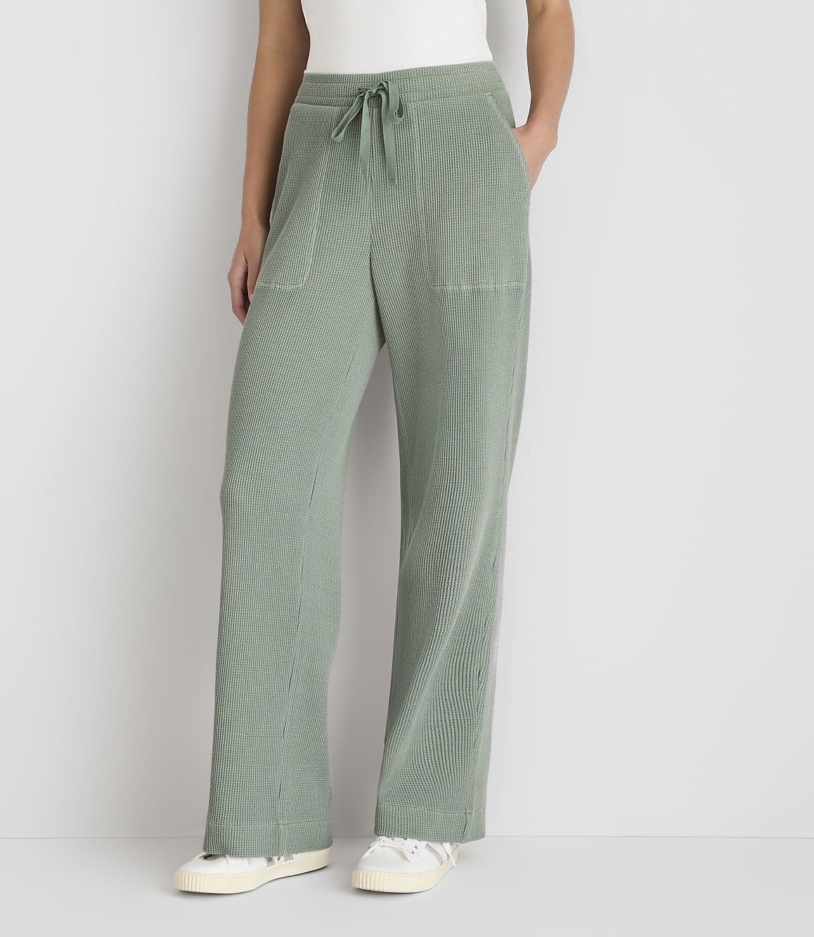 A person stands in front of a plain white background, wearing loose-fitting, light green, ribbed pants with white sneakers.