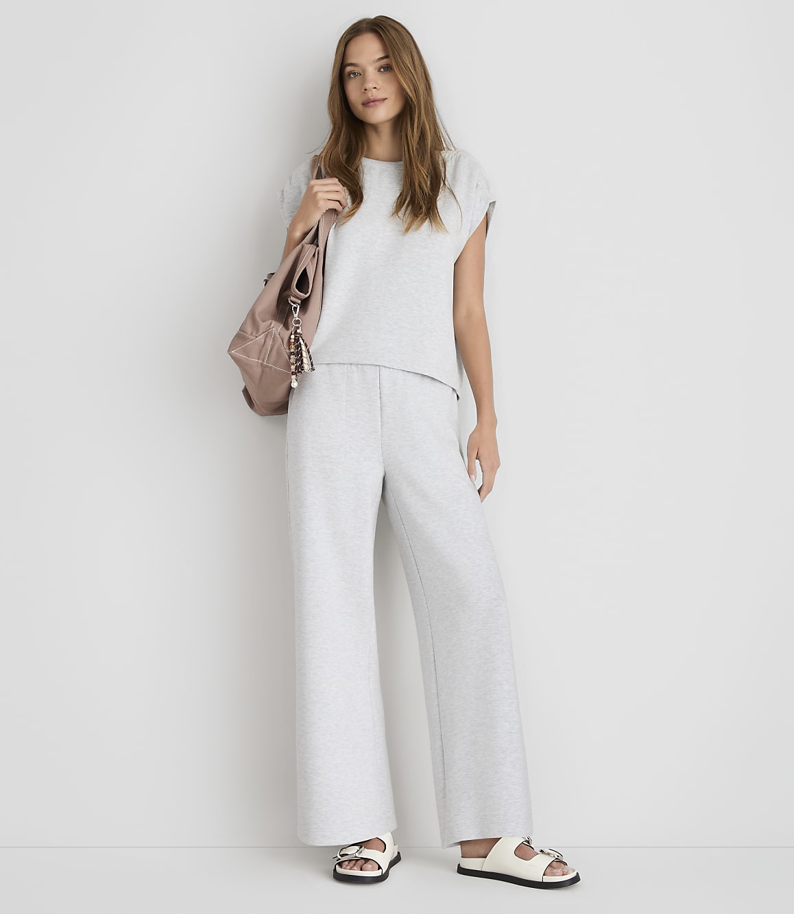 A young woman in a grey loungewear set and white sandals stands against a plain white background, with a pink backpack slung over her shoulder.