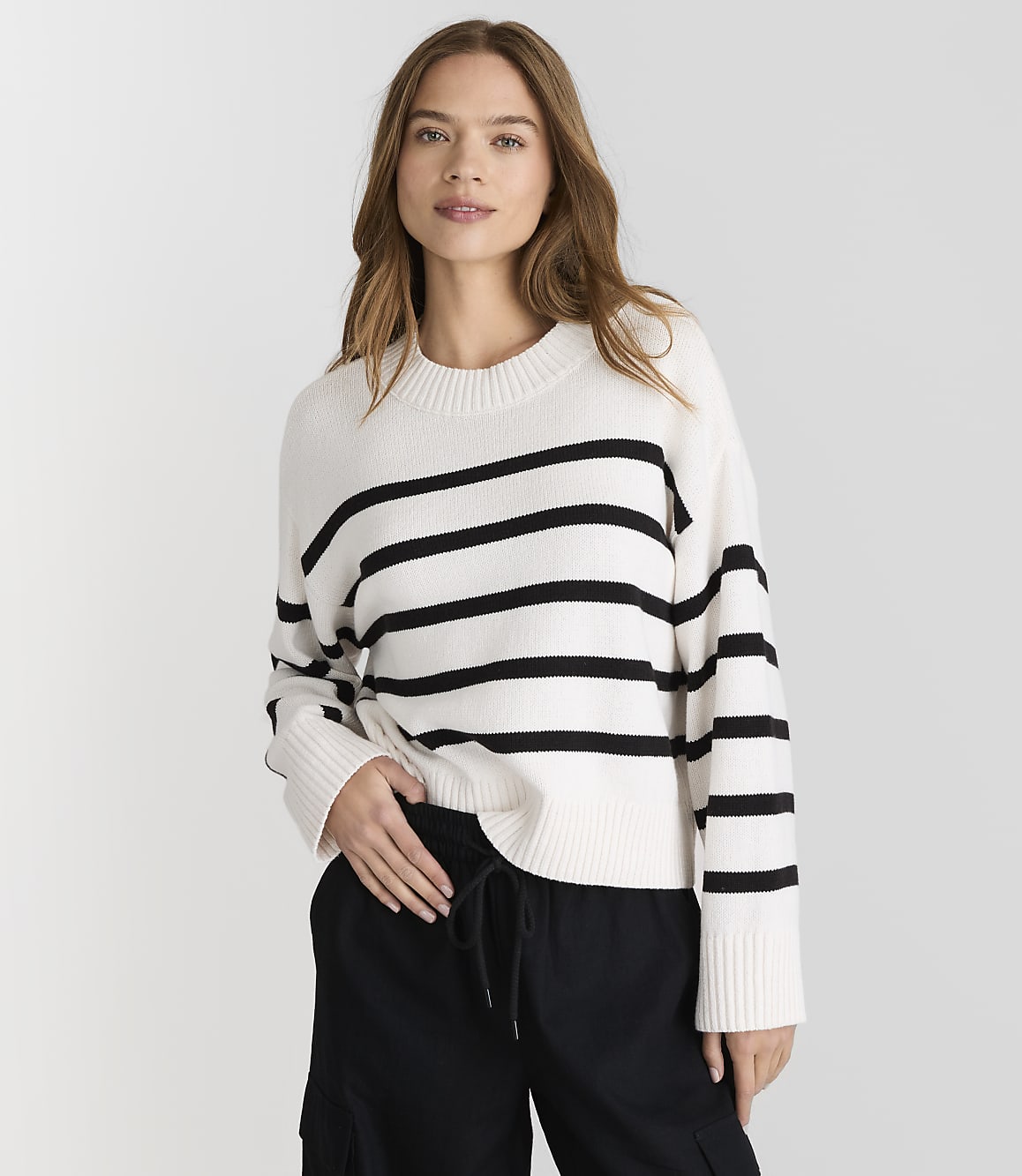 A woman in a striped sweater and black pants stands against a plain white background.