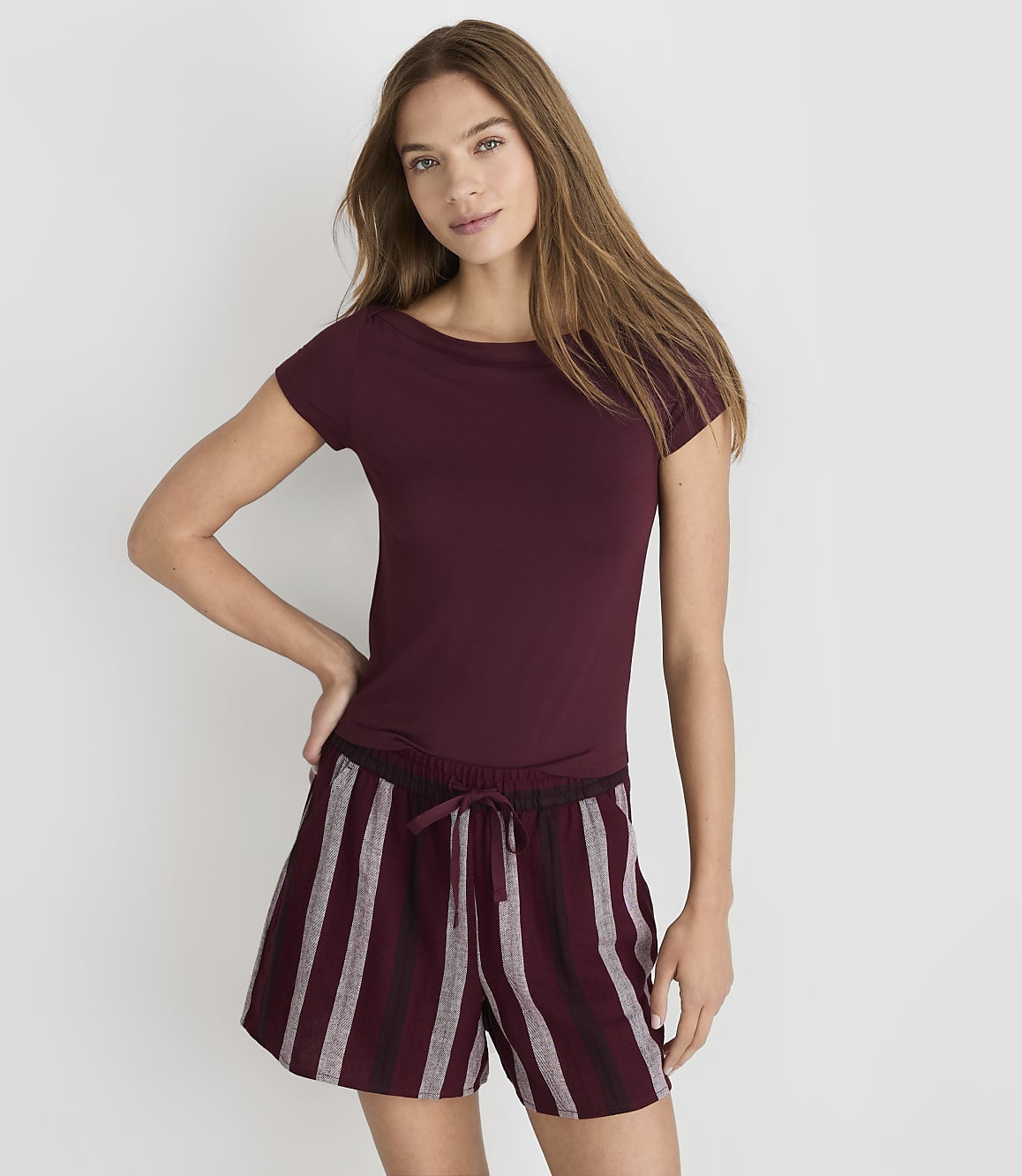 A woman in a maroon t-shirt and striped shorts stands against a plain white background.