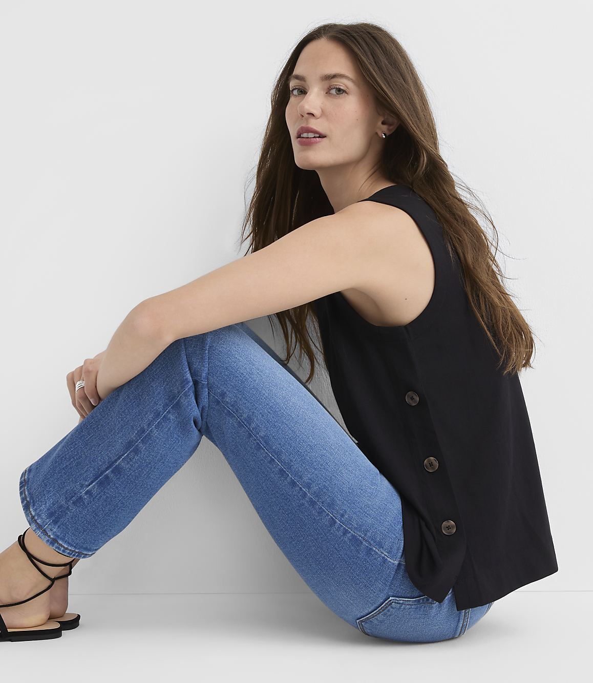 A woman in a black sleeveless top and blue jeans sits against a plain white background.