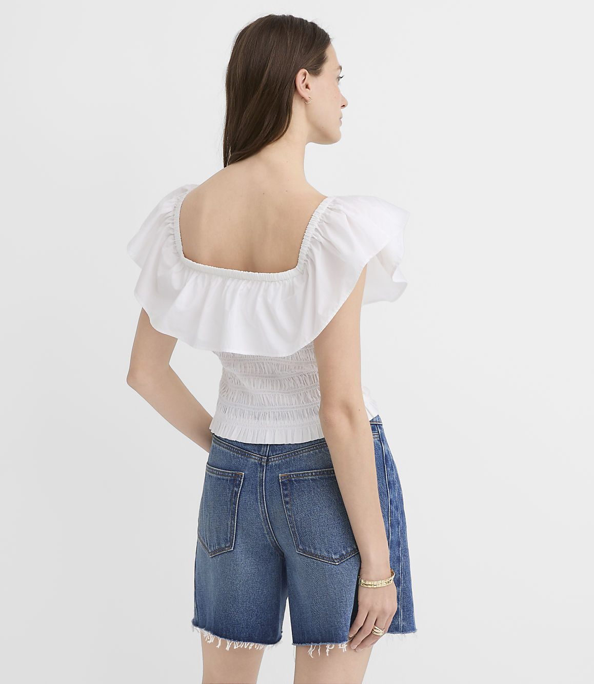 A woman in a white ruffled top and denim shorts stands against a plain white background.
