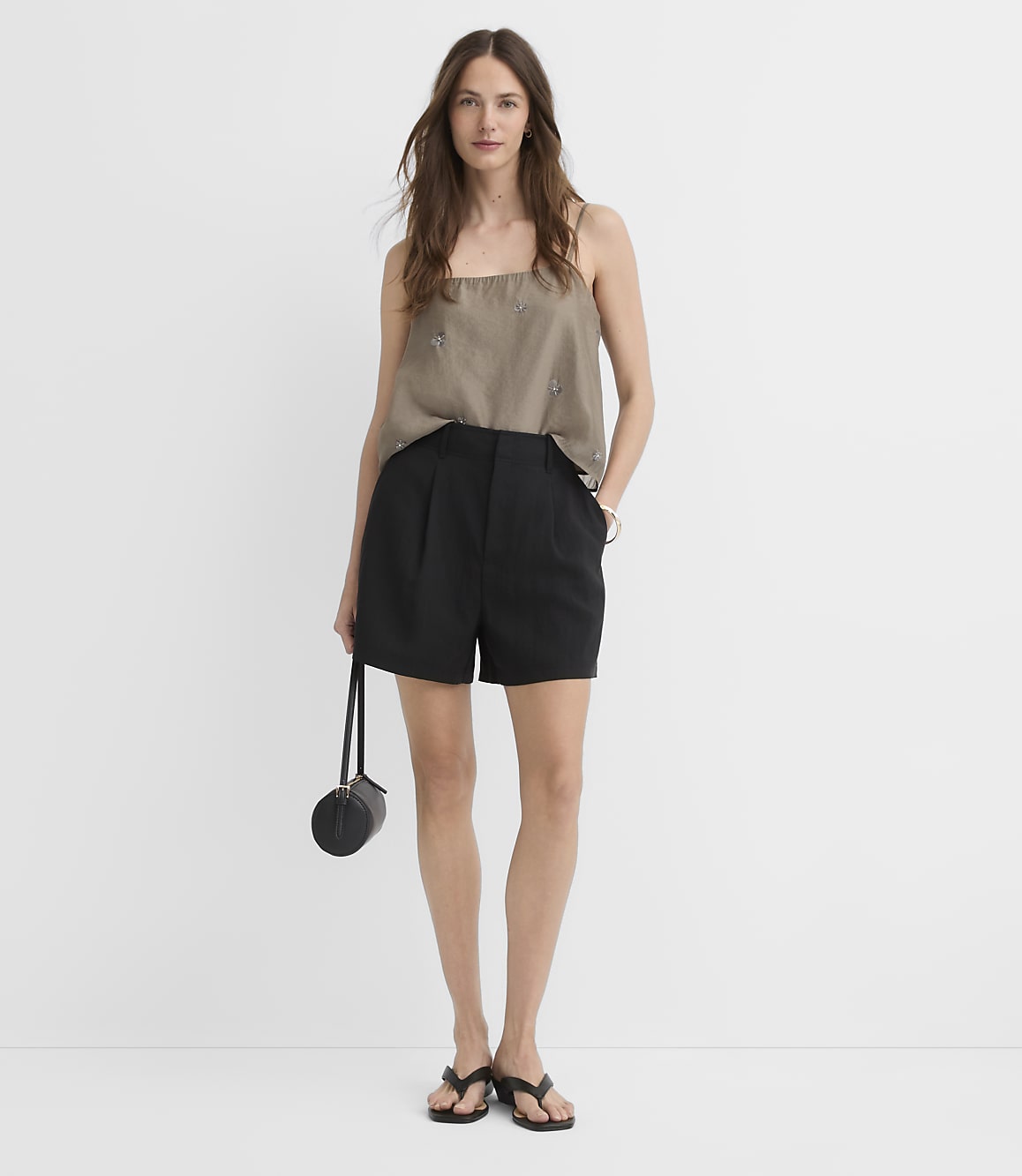 A woman in a taupe camisole and black shorts stands against a plain white background, holding a small black purse.