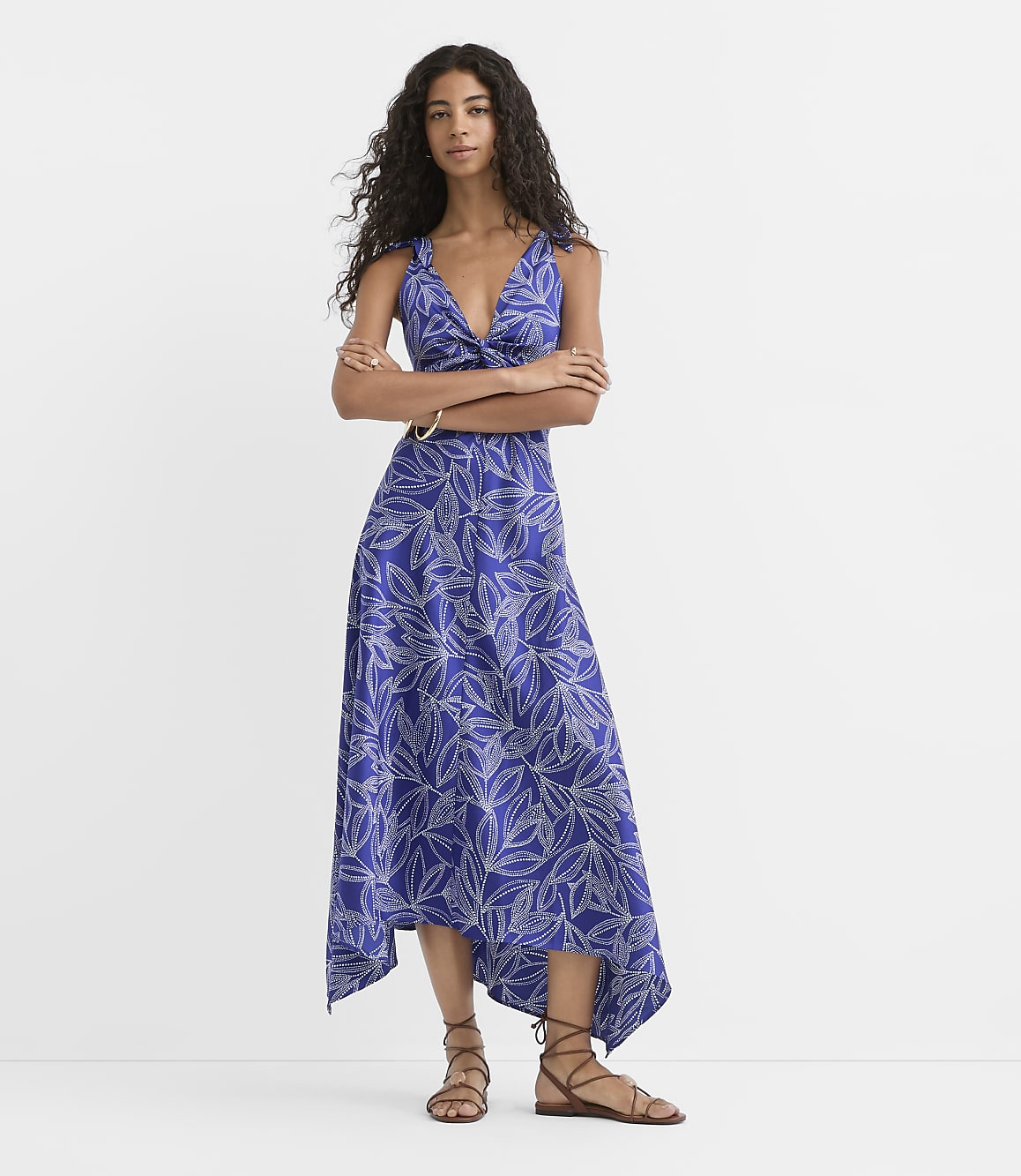 A woman in a blue floral dress stands with her arms crossed against a plain white background.