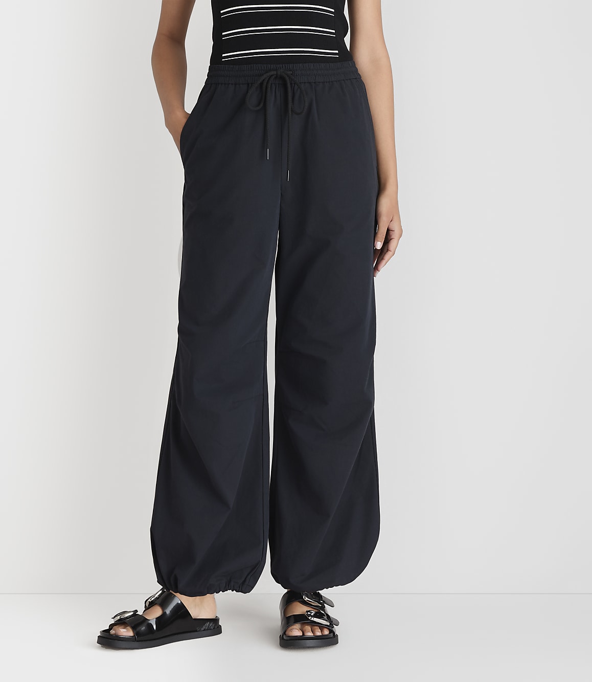 A person stands in black balloon pants and sandals against a plain white background.