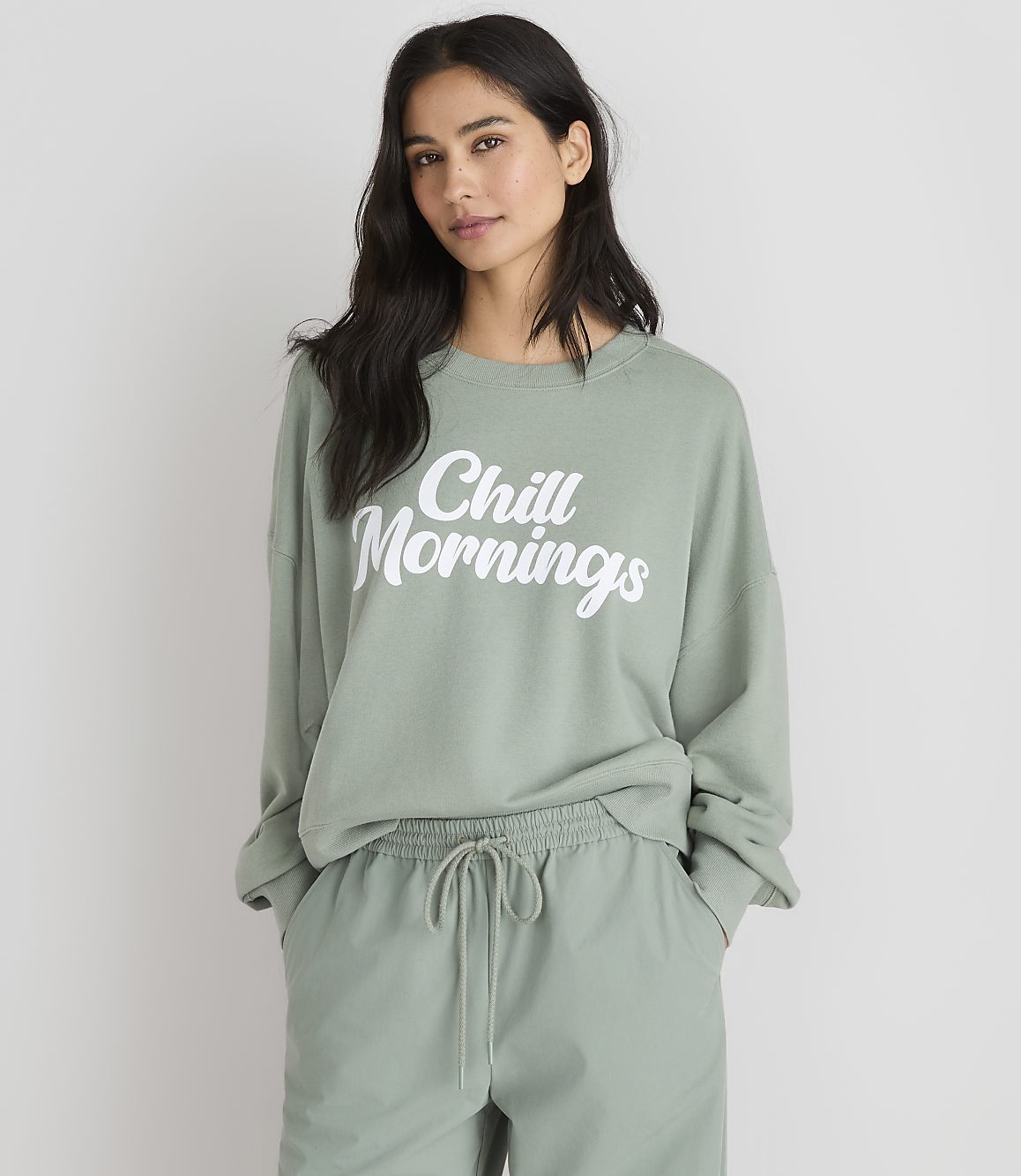 A woman in a light green sweatshirt with "Chill Mornings" printed on it stands against a plain white background.