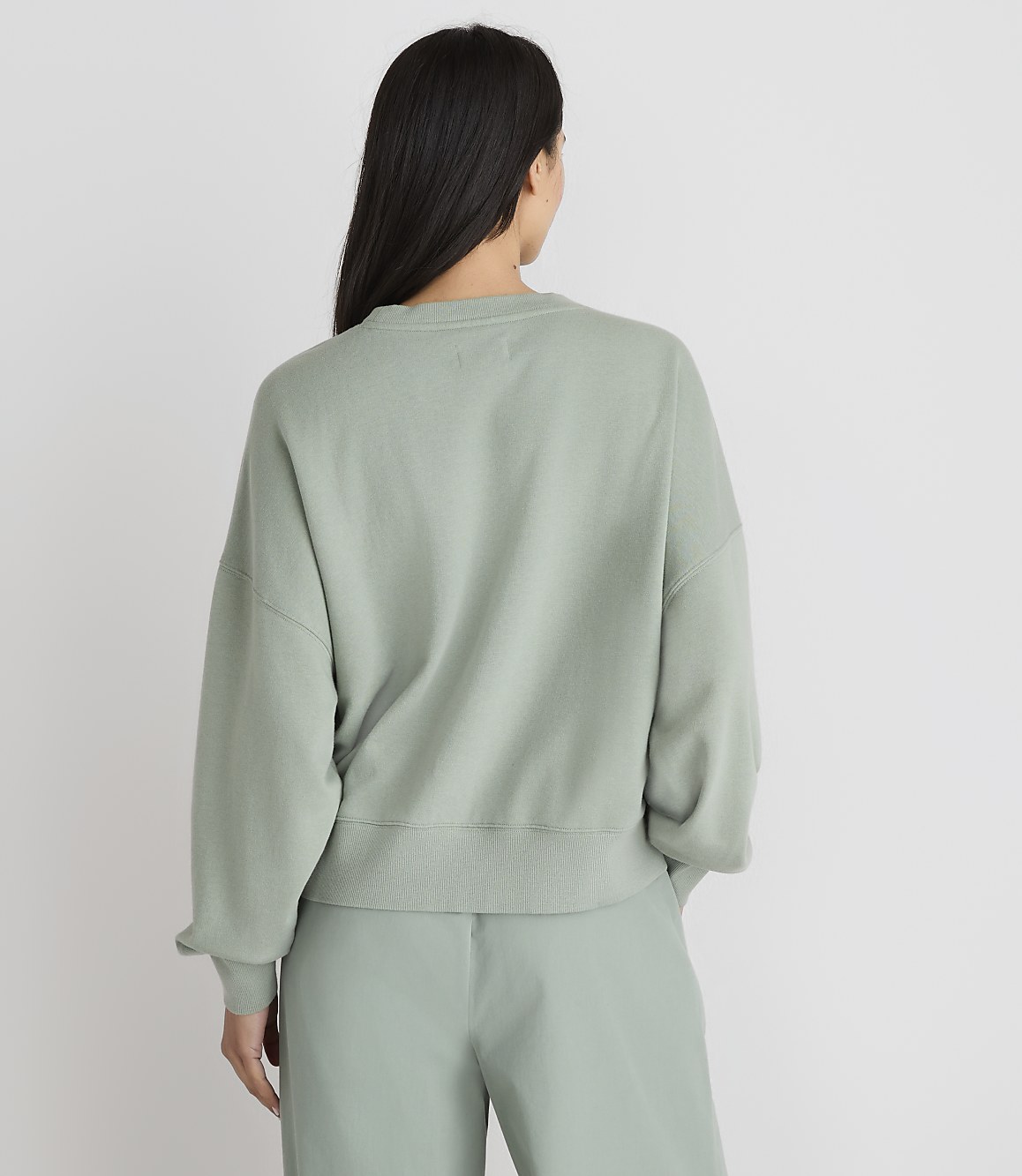 A woman in a light green sweatshirt and pants stands with her back to the camera against a plain white background.