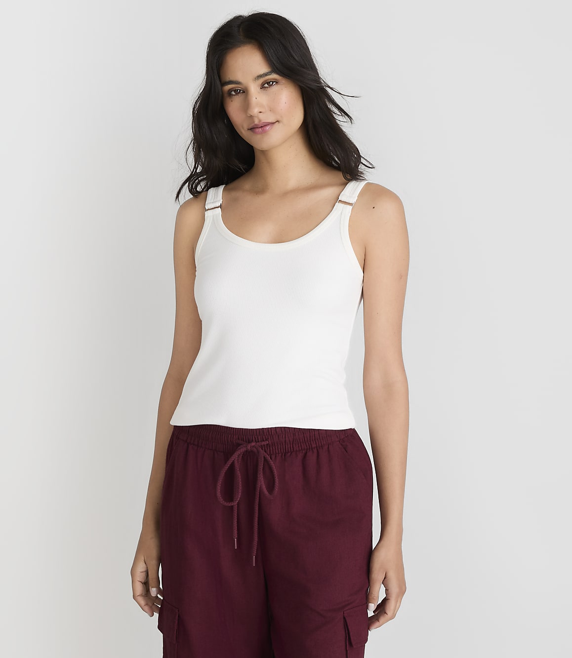 A woman in a white tank top and maroon pants stands against a plain white background.