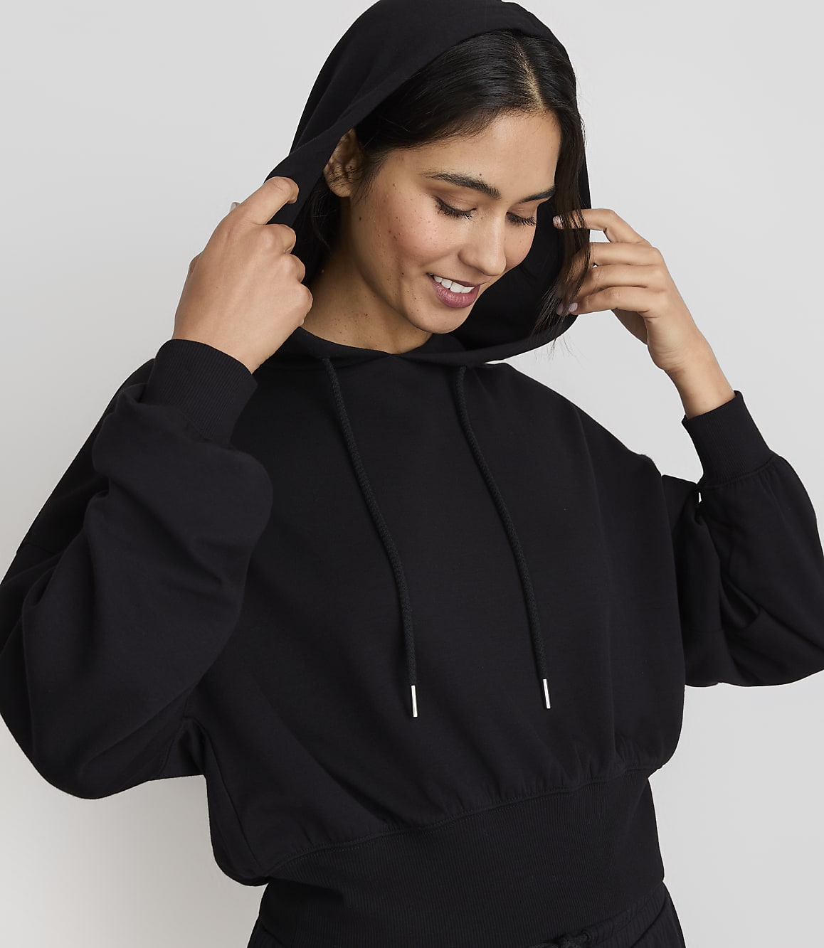A smiling woman in a black hoodie adjusts the hood against a plain gray background.