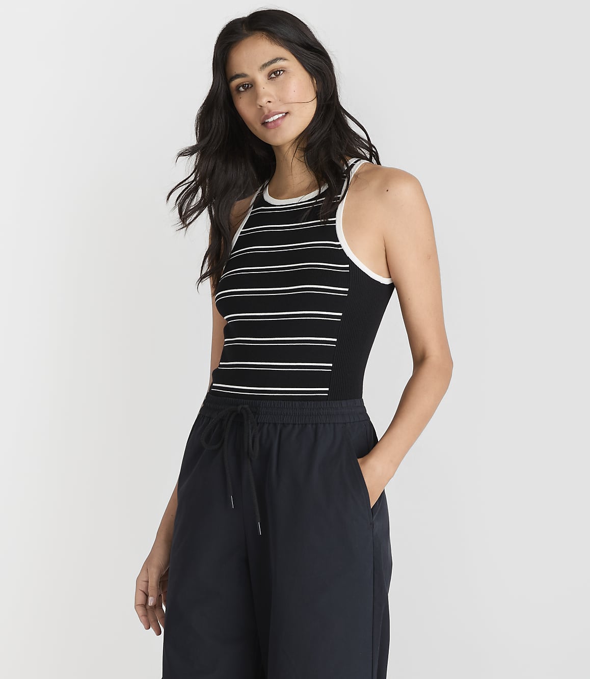 A woman in a striped tank top and dark pants stands against a plain white background.