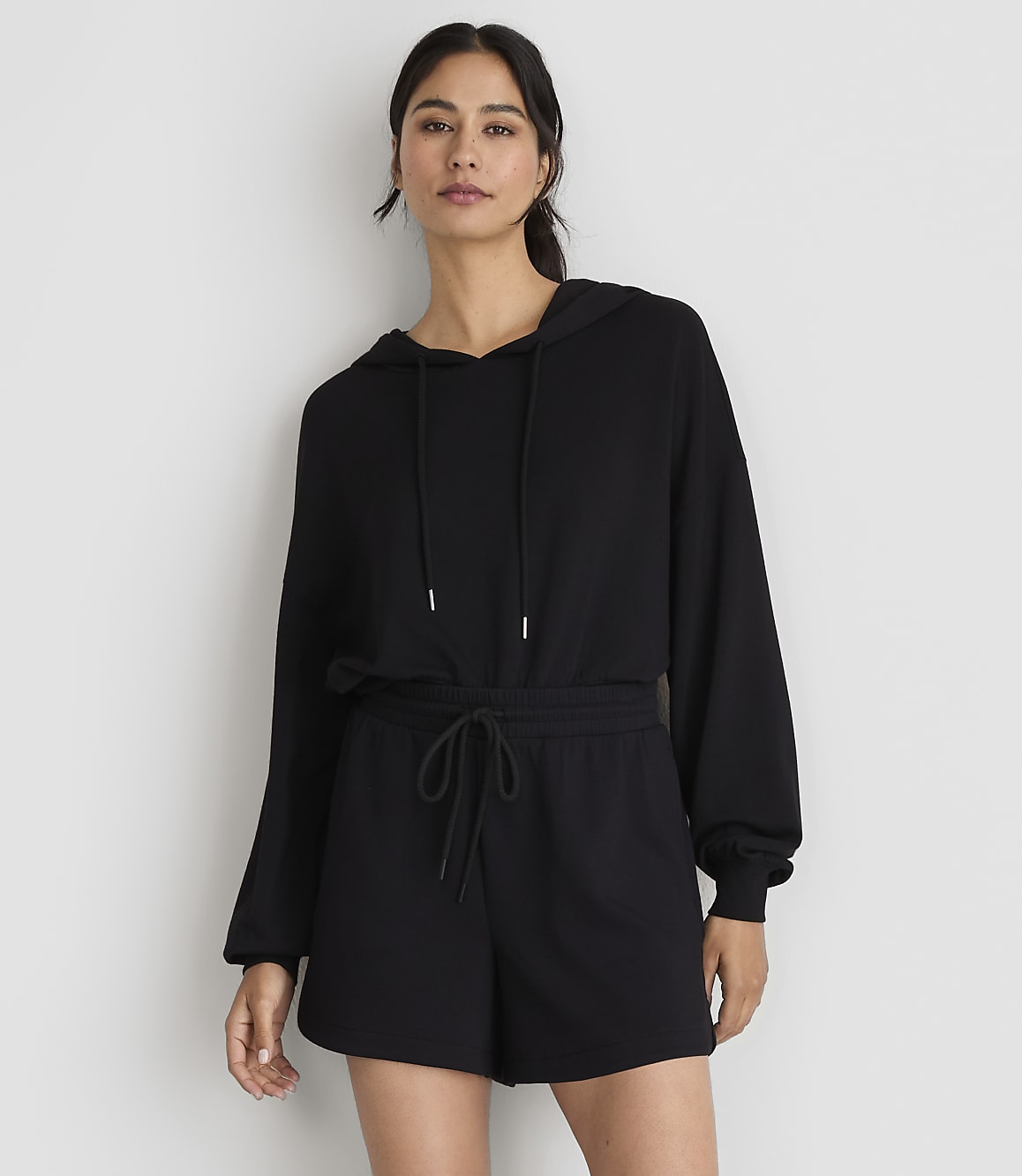 A woman in a black hoodie and shorts stands against a plain white background.