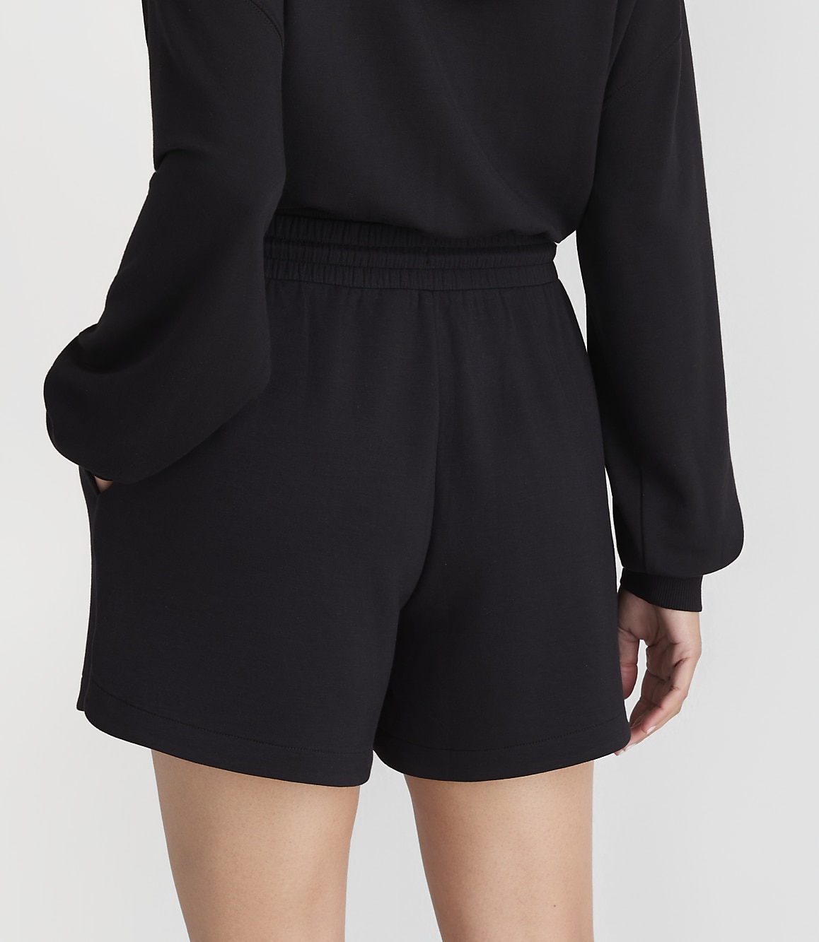 A person wearing black shorts and a black sweatshirt stands against a plain gray background.