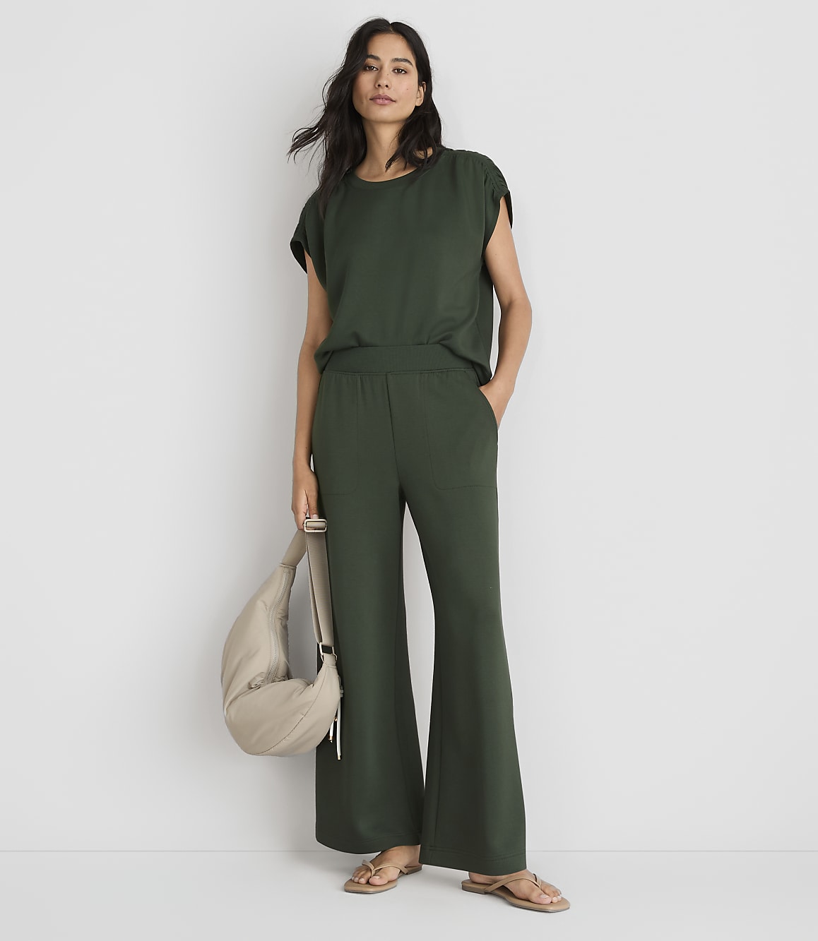 A woman in a green top and wide-leg pants stands against a plain white background, holding a beige bag.