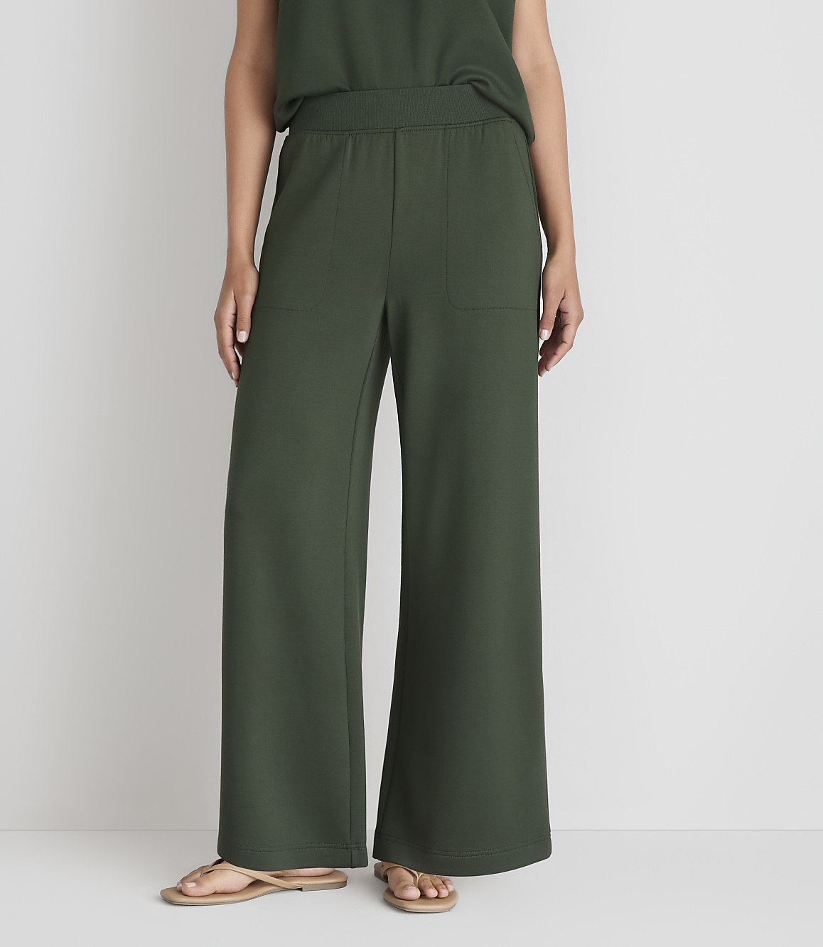 A person wearing dark green wide-leg pants and sandals stands against a plain white background.