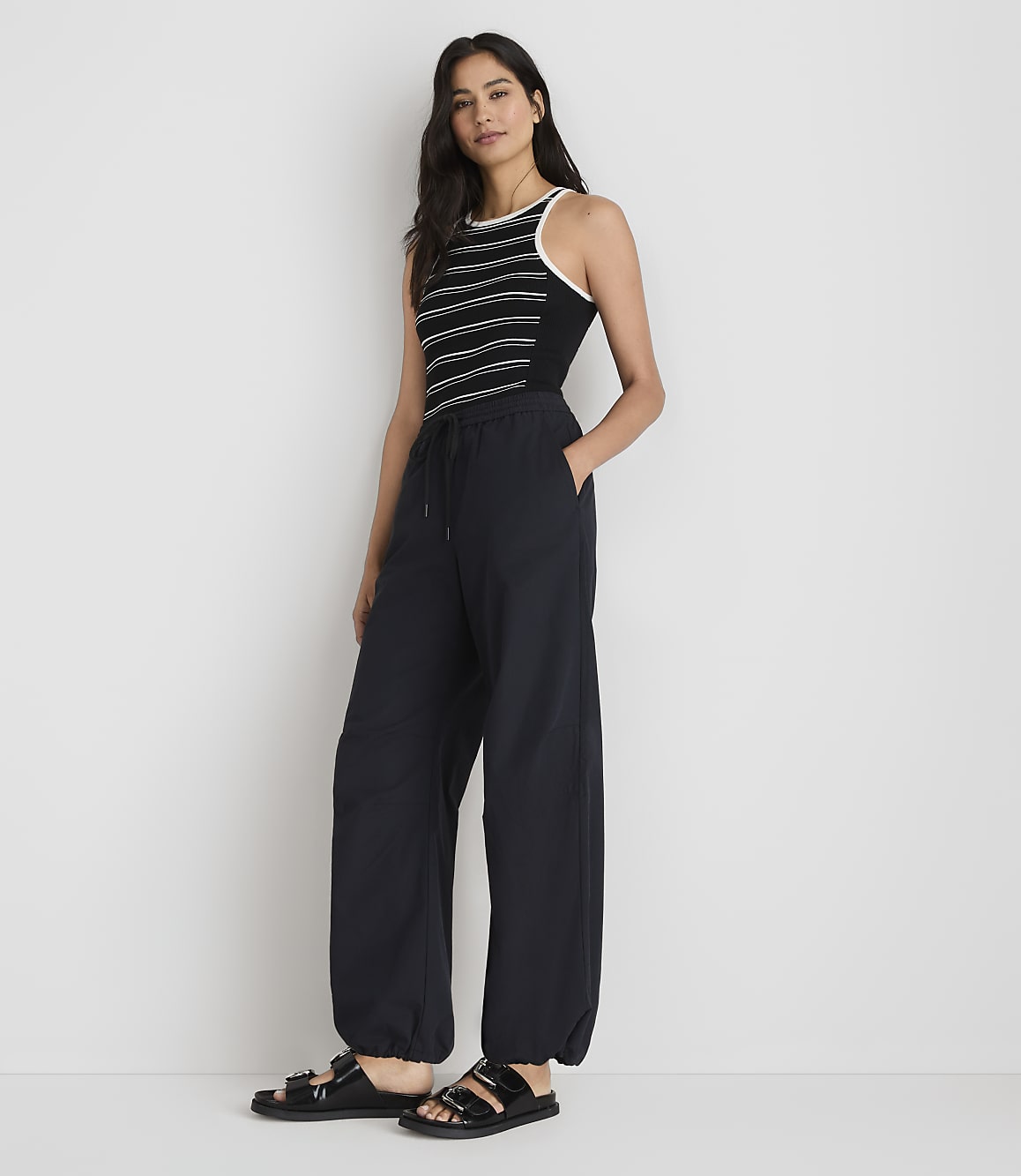 A woman in a striped tank top and baggy pants stands against a plain white background.