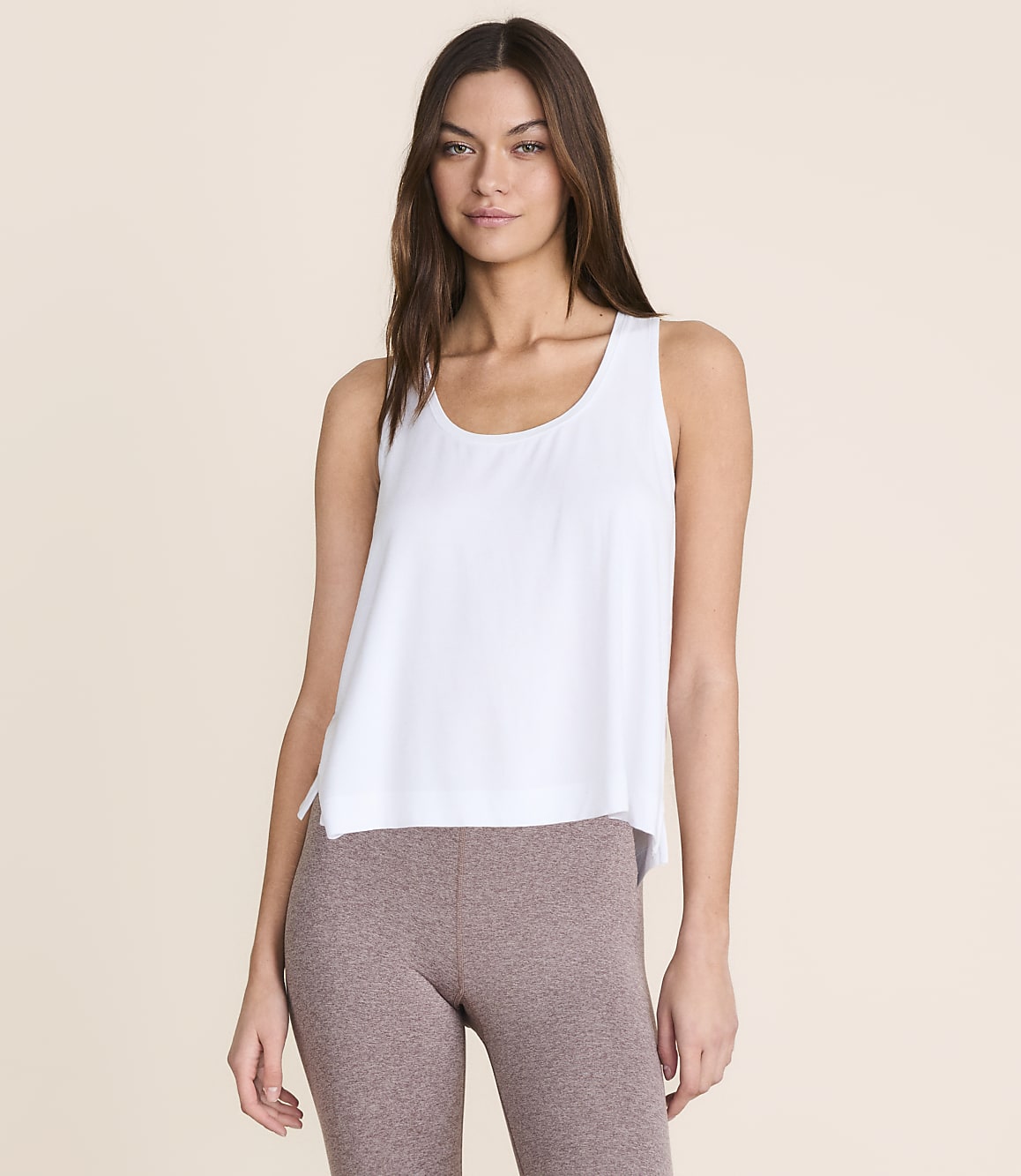A woman in a white tank top and brown leggings stands against a plain, light peach background.