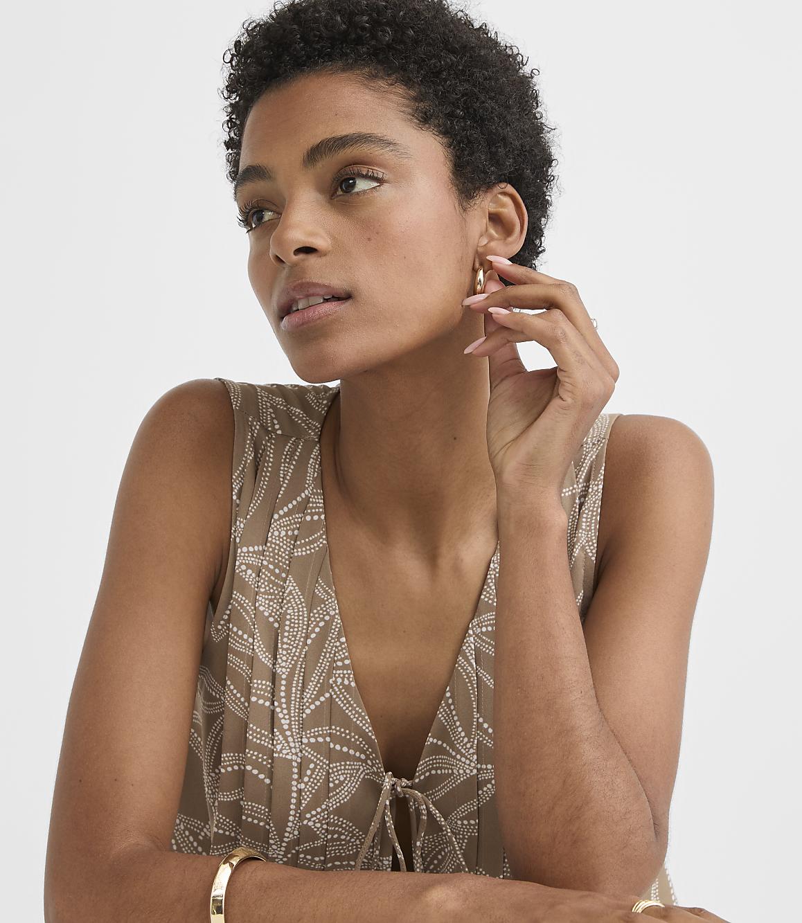 A woman with short, dark hair and a patterned top touches her ear while a plain background surrounds her.