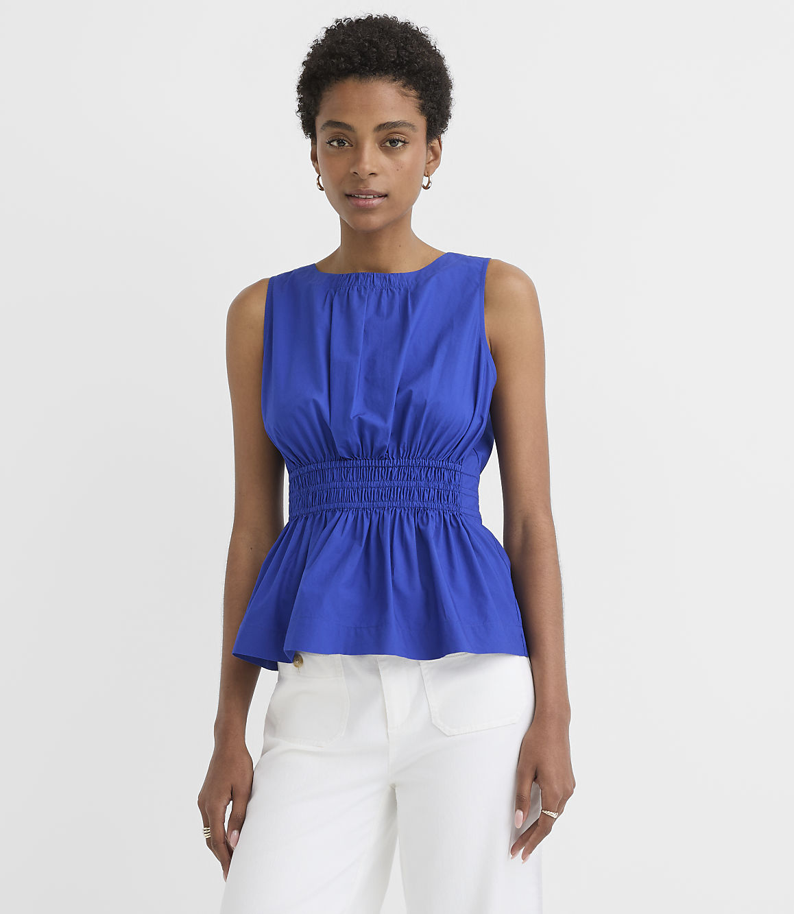 A woman in a blue peplum top and white pants stands against a plain white background.