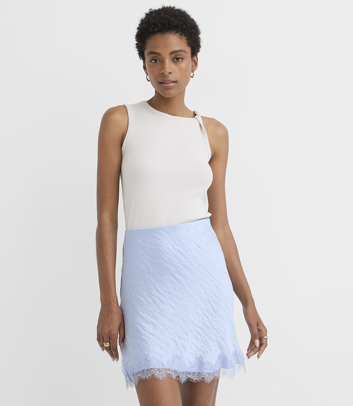 A woman in a light gray sleeveless top and a light blue textured skirt with lace trim stands against a plain white background.