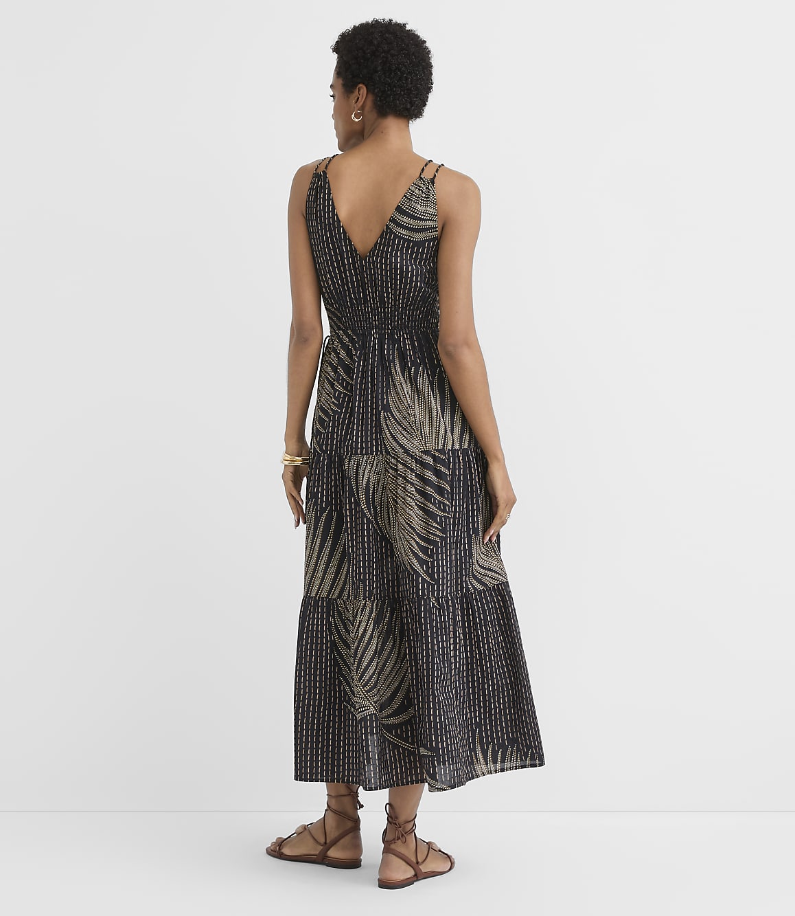 A woman in a black and gold palm-print tiered maxi dress stands against a plain white background.