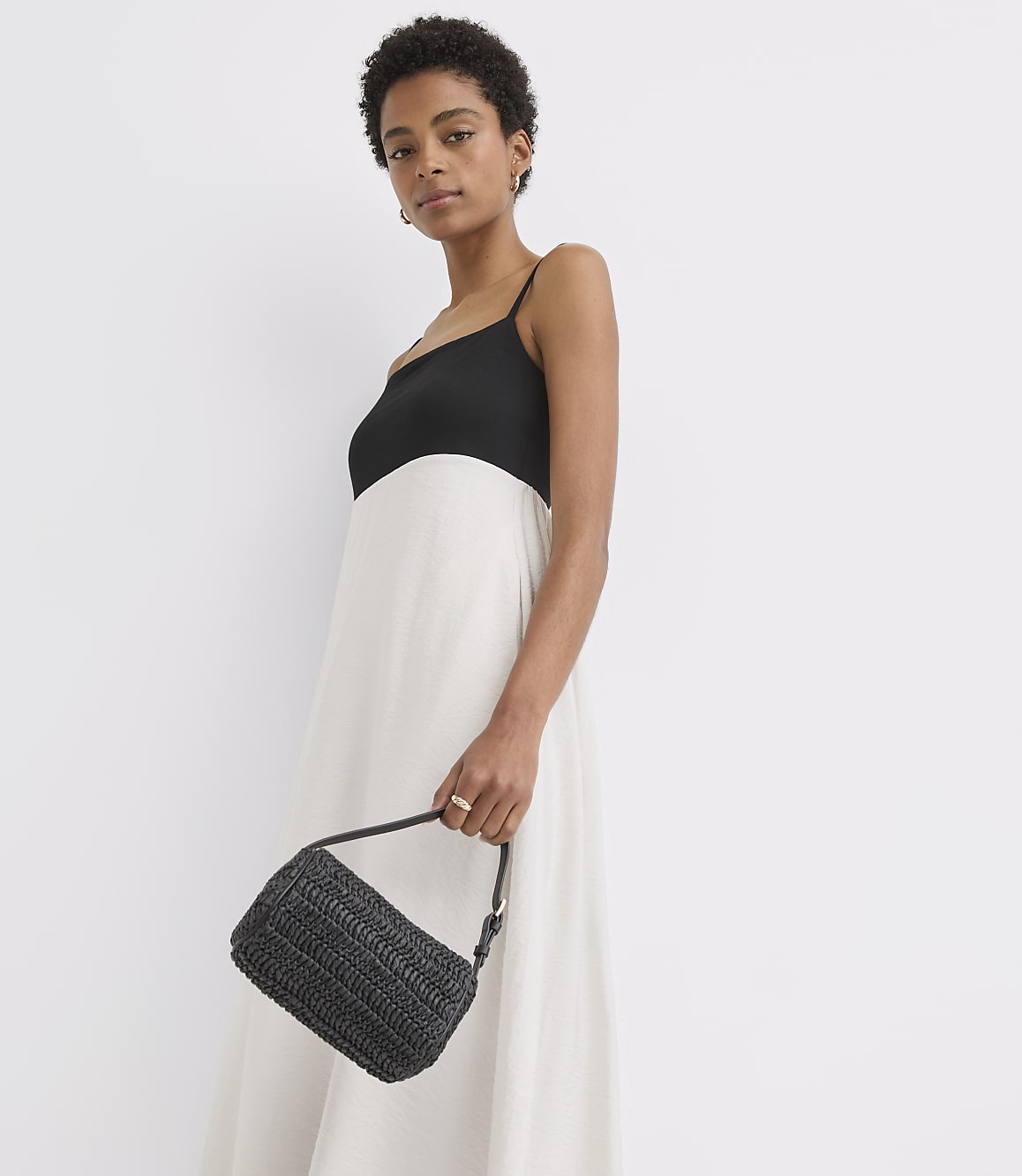 A woman in a white and black dress holds a black woven purse against a plain white background.
