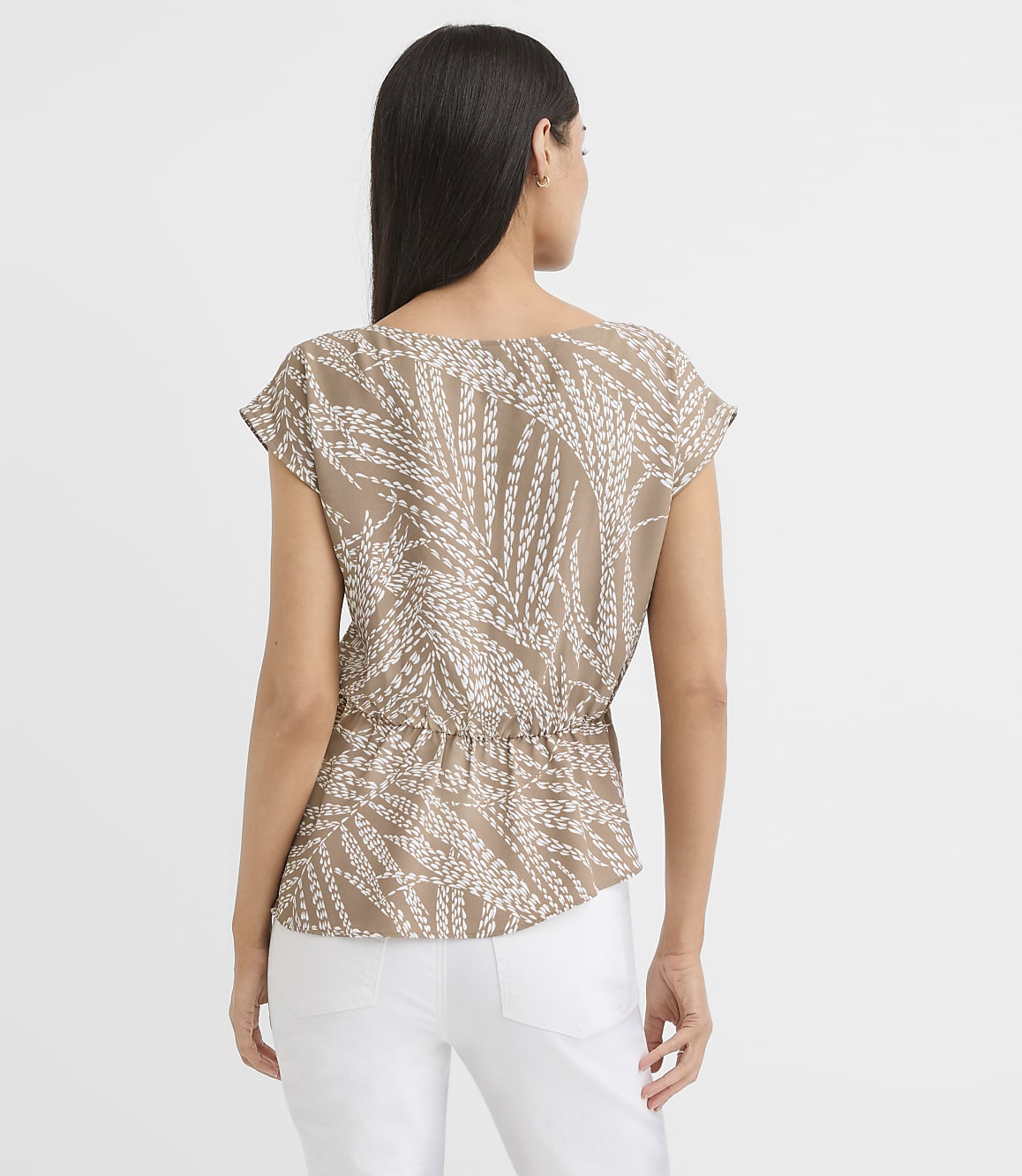 A woman in a tan, leaf-patterned blouse and white pants stands against a plain white background.