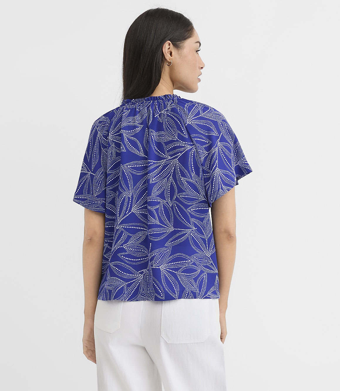 A woman in a blue, leaf-patterned top and white pants stands against a plain white background.