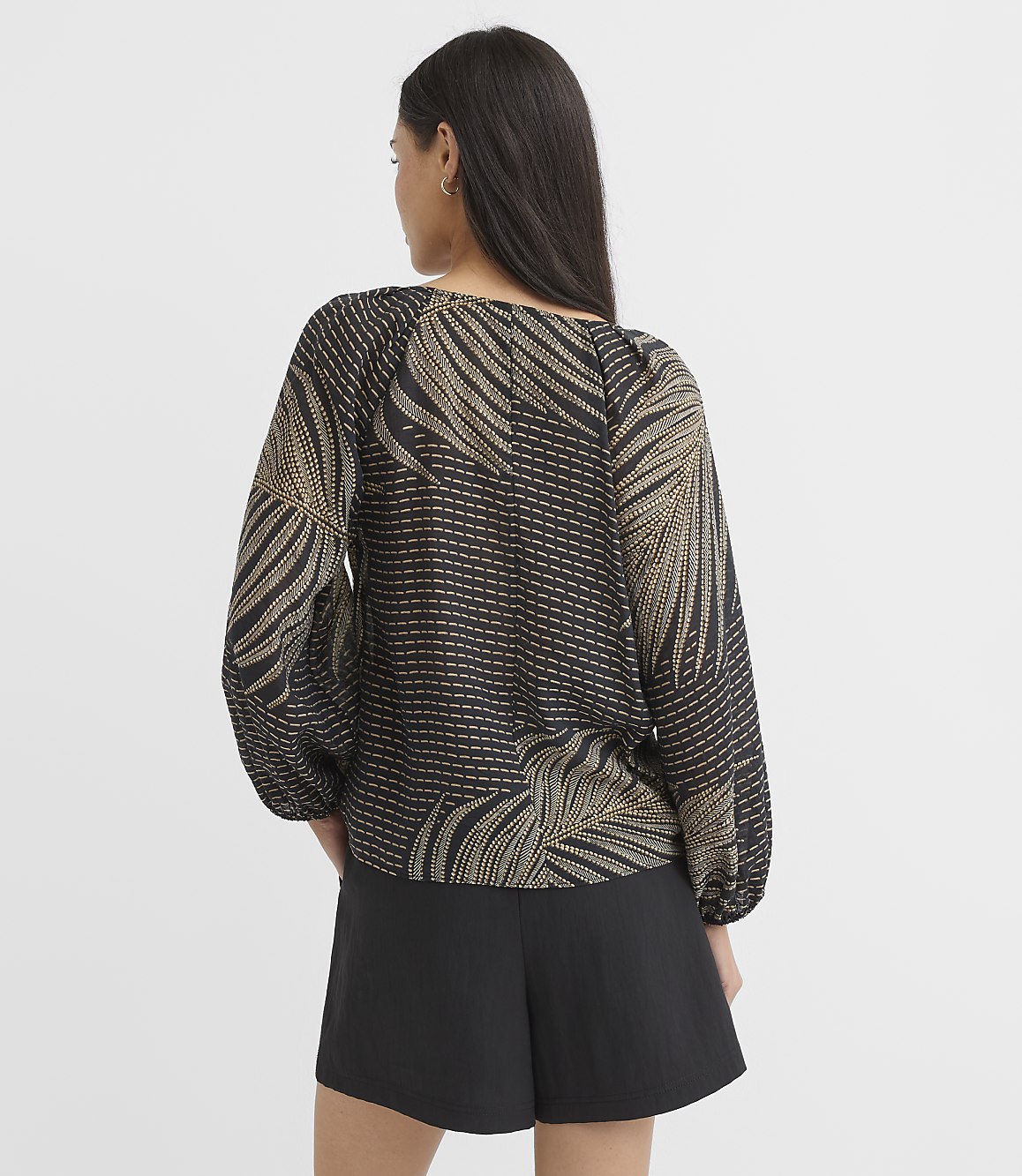 A woman in a black and gold patterned blouse and black shorts stands against a plain white background.