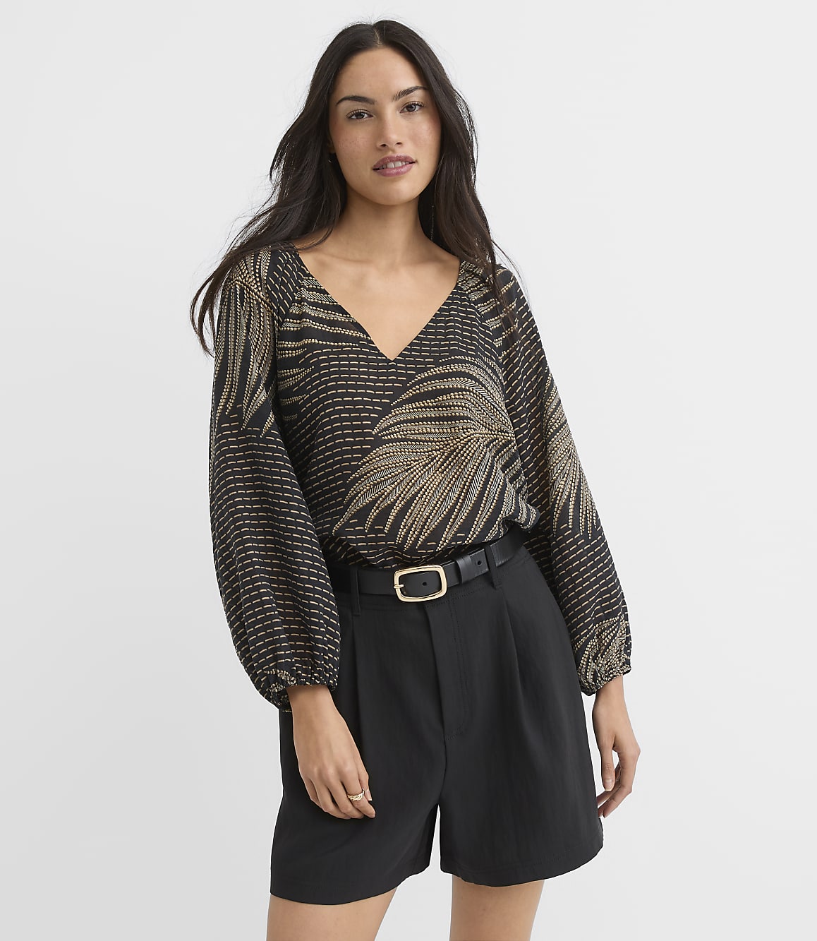 A woman in a patterned blouse and black shorts stands against a plain white background.