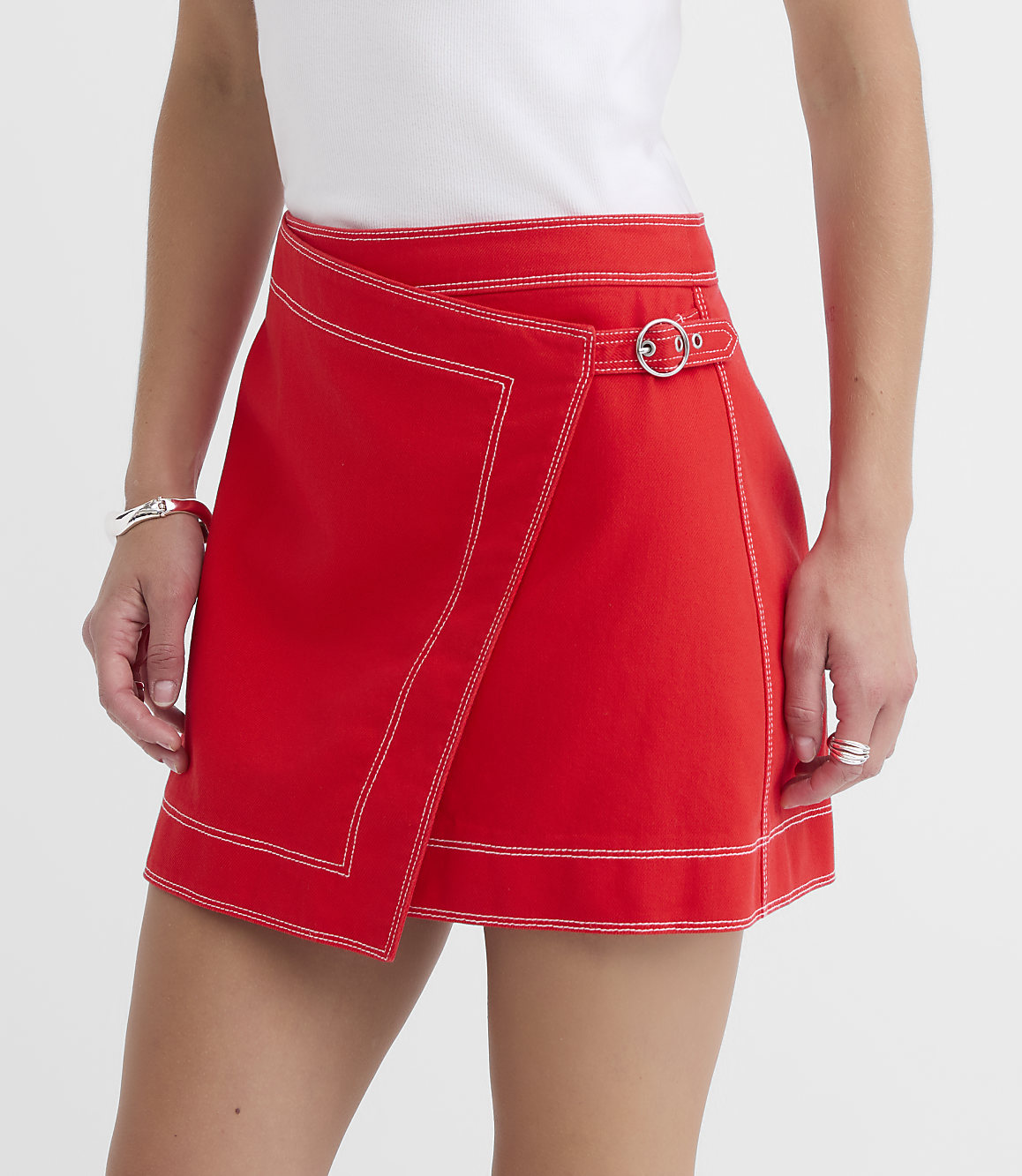 A person models a red wrap skirt with white stitching and a silver buckle in the foreground, against a plain white background.