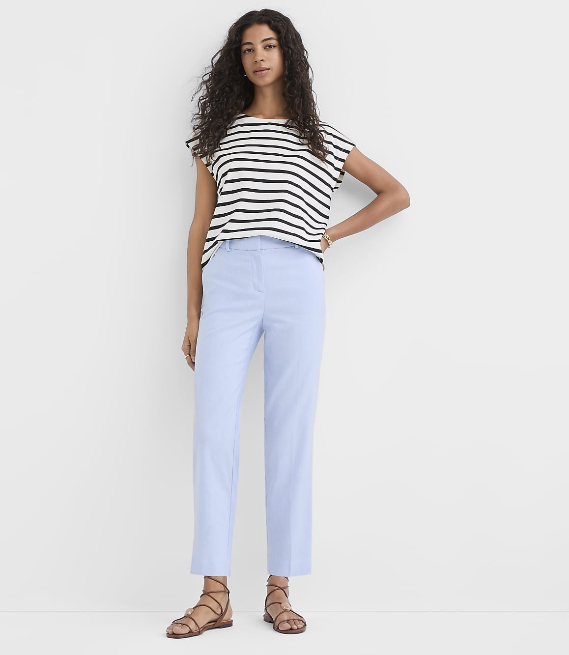 A woman in a striped shirt and light blue pants stands against a plain white background.