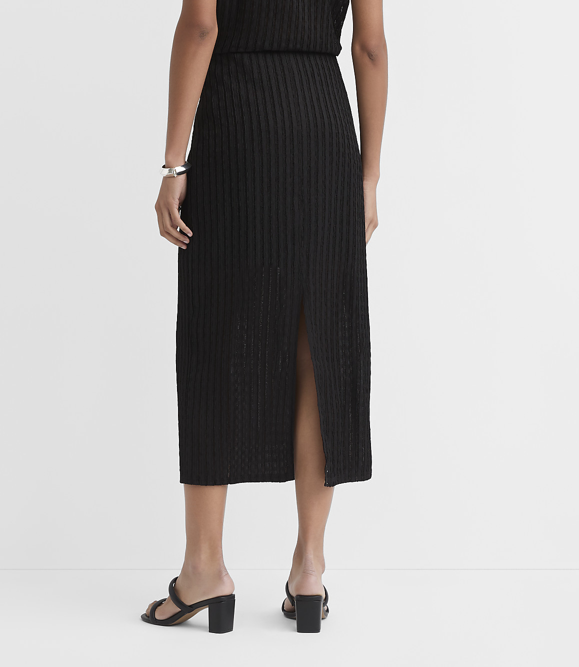 A person wearing a black ribbed midi skirt with a slit and black block heels stands against a plain white background.