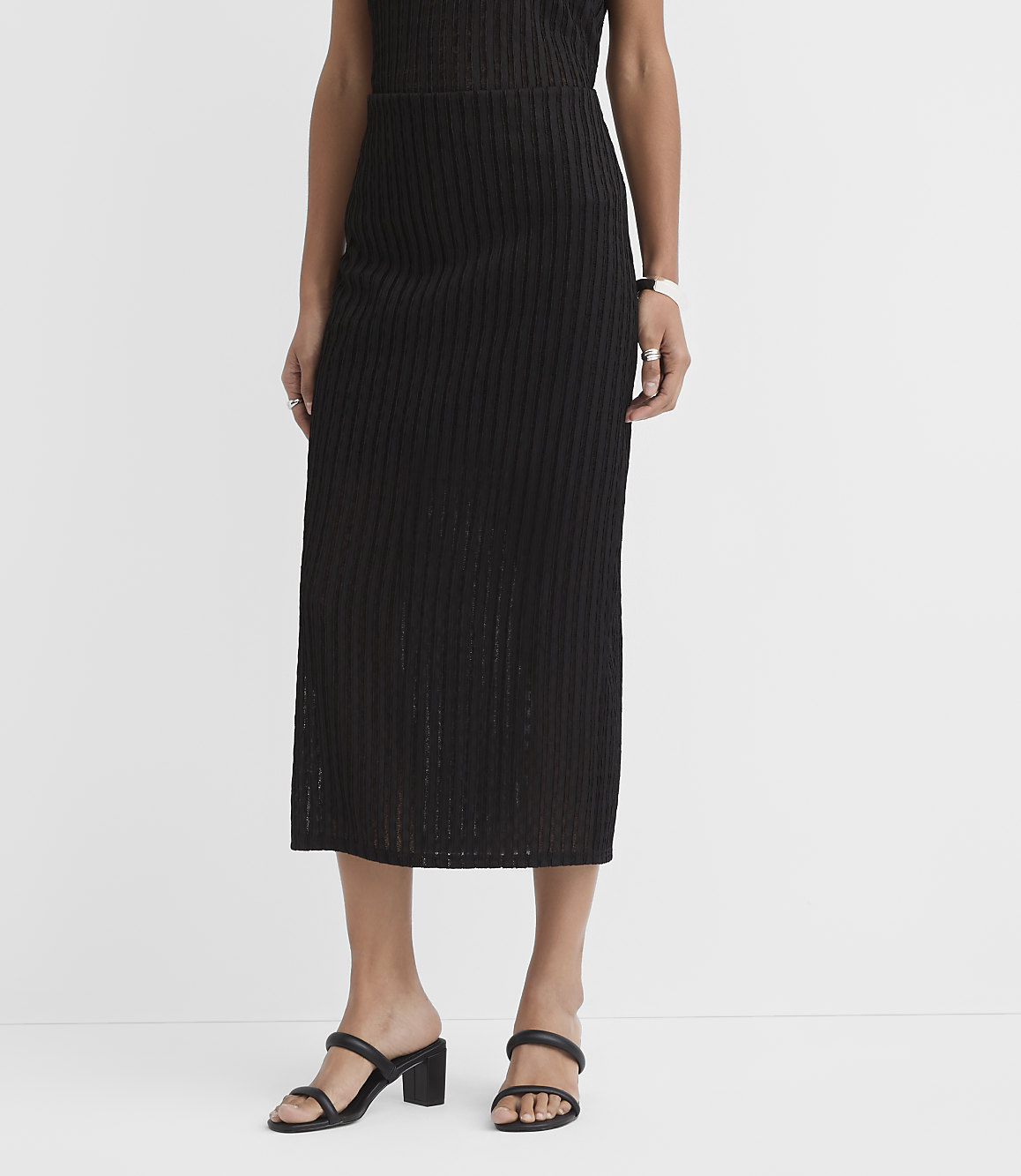 A person wearing a black ribbed midi skirt and black block-heeled sandals stands against a plain white background.
