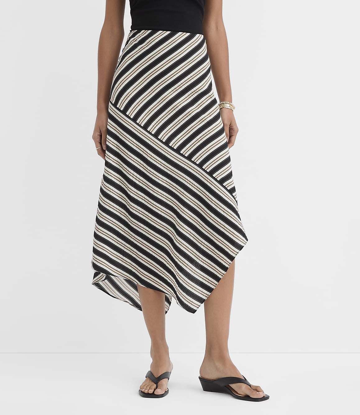 A woman models a black, white, and tan striped asymmetrical midi skirt with black wedge sandals against a plain white background.