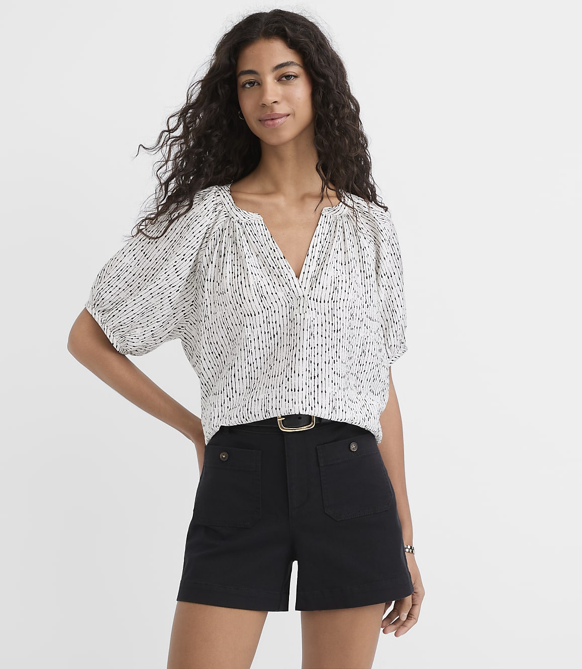 A woman with curly dark hair stands in the foreground wearing a patterned white blouse and black shorts, against a plain white background.