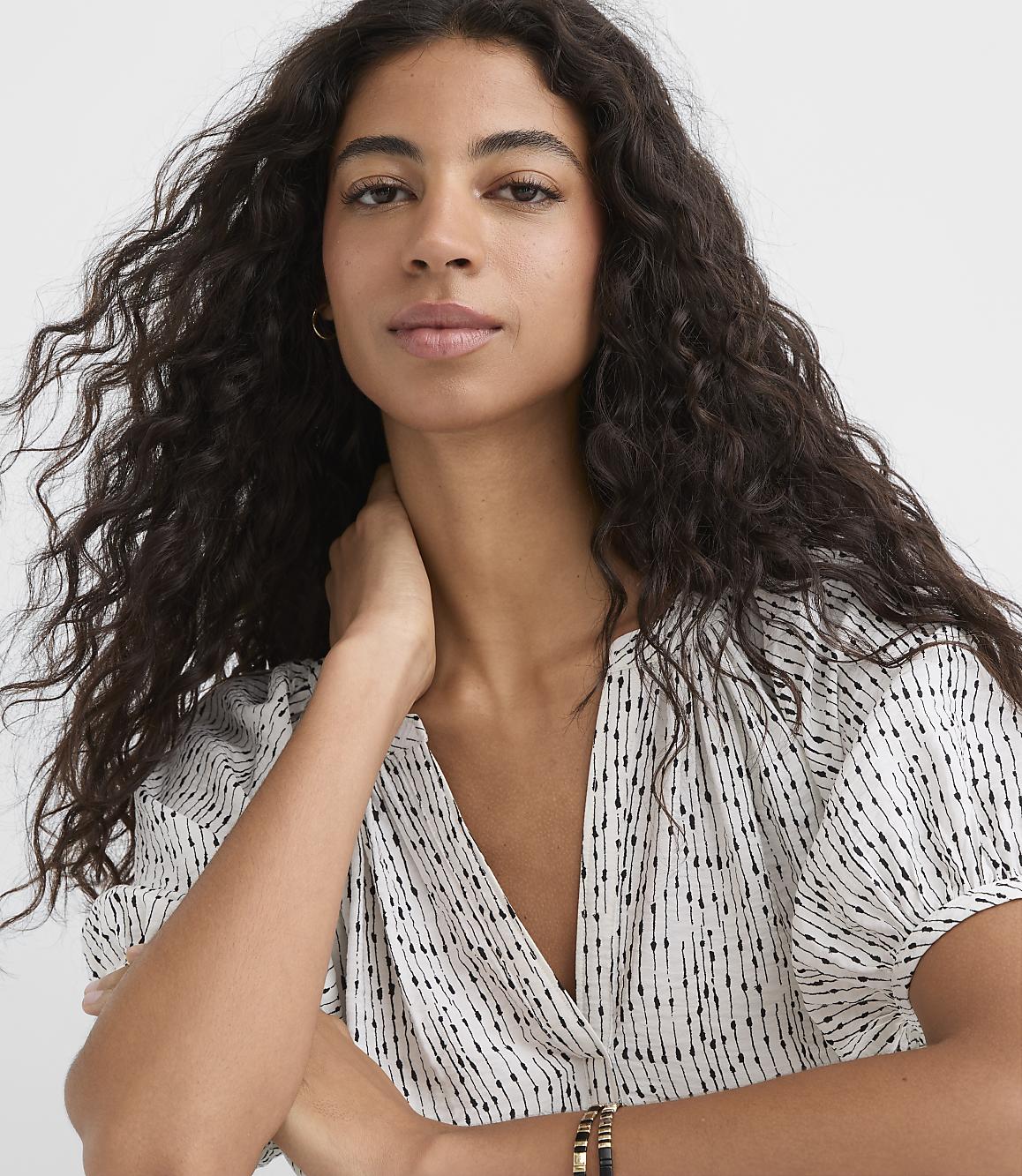 A woman with long, dark, curly hair and a patterned top is shown against a plain white background.