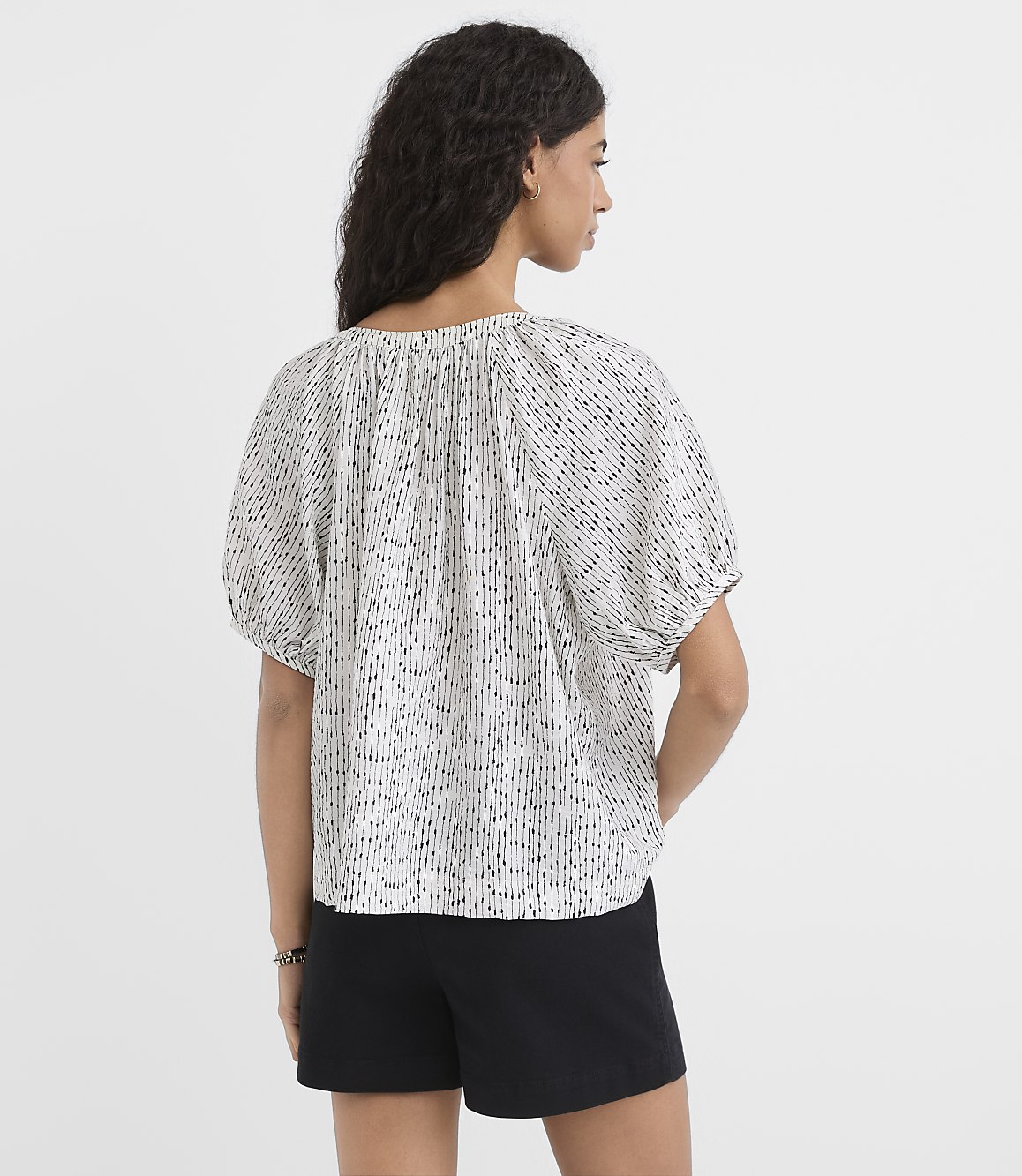 A woman in a patterned blouse and black shorts stands against a plain white background.