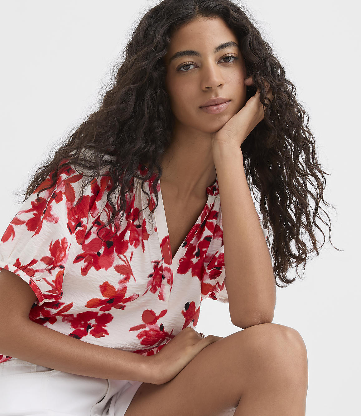 A young woman with long, dark, curly hair rests her chin on her hand, wearing a white top with a red floral print, against a plain white background.