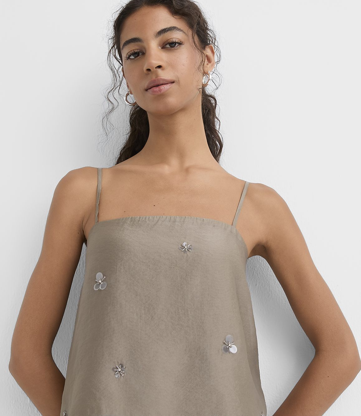 A woman in a taupe camisole with floral embellishments stands against a plain white background.