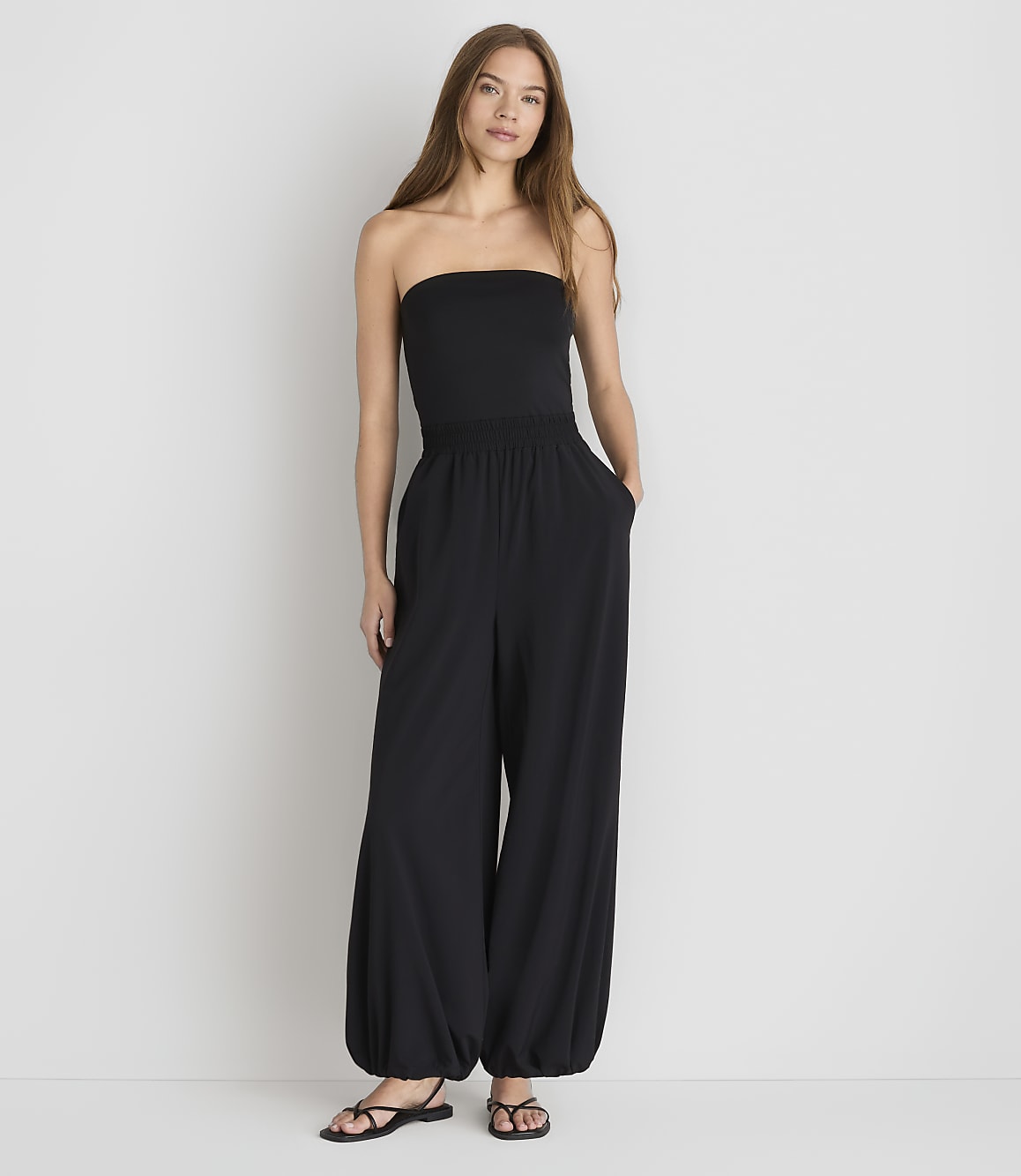 A woman in a black strapless jumpsuit stands against a plain white background.