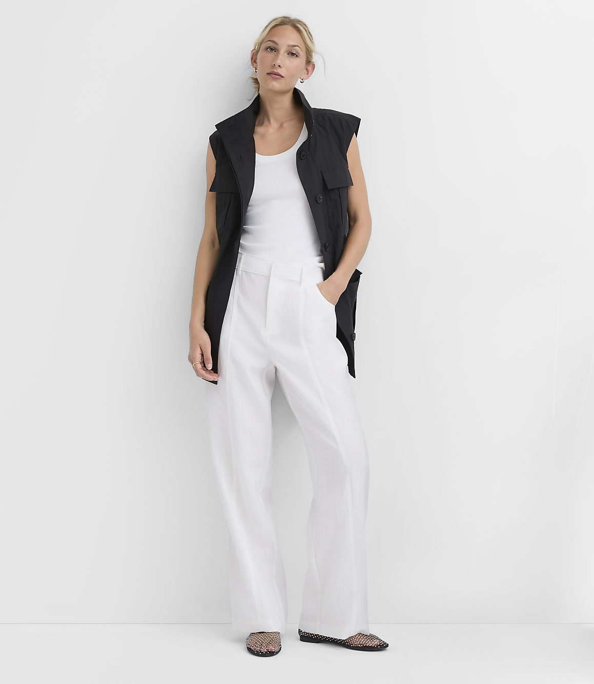 A woman in a white tank top, black vest, and white wide-leg pants stands against a plain white background.